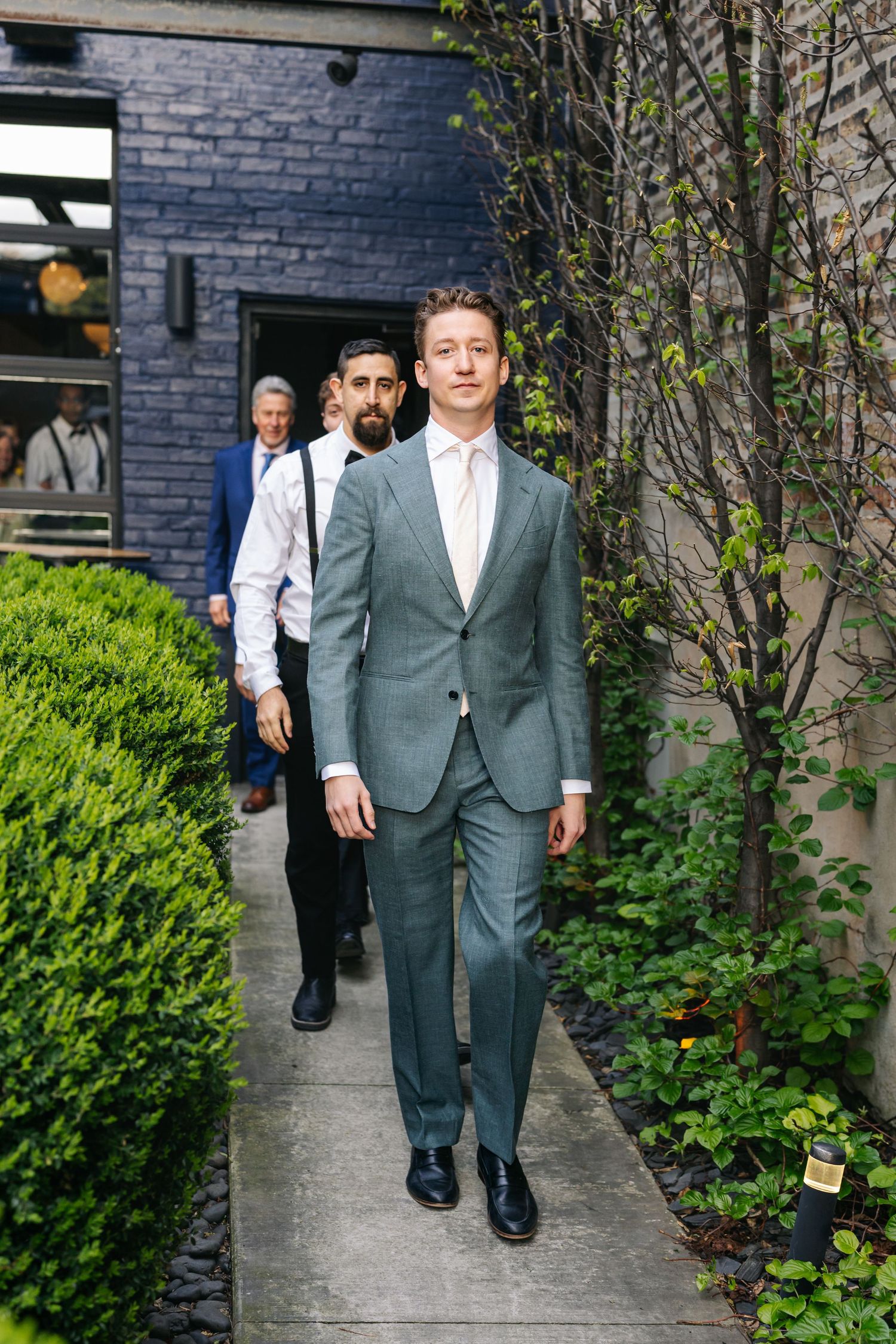 Groom in teal suit walks down garden path with groomsmen in suspenders following behind.