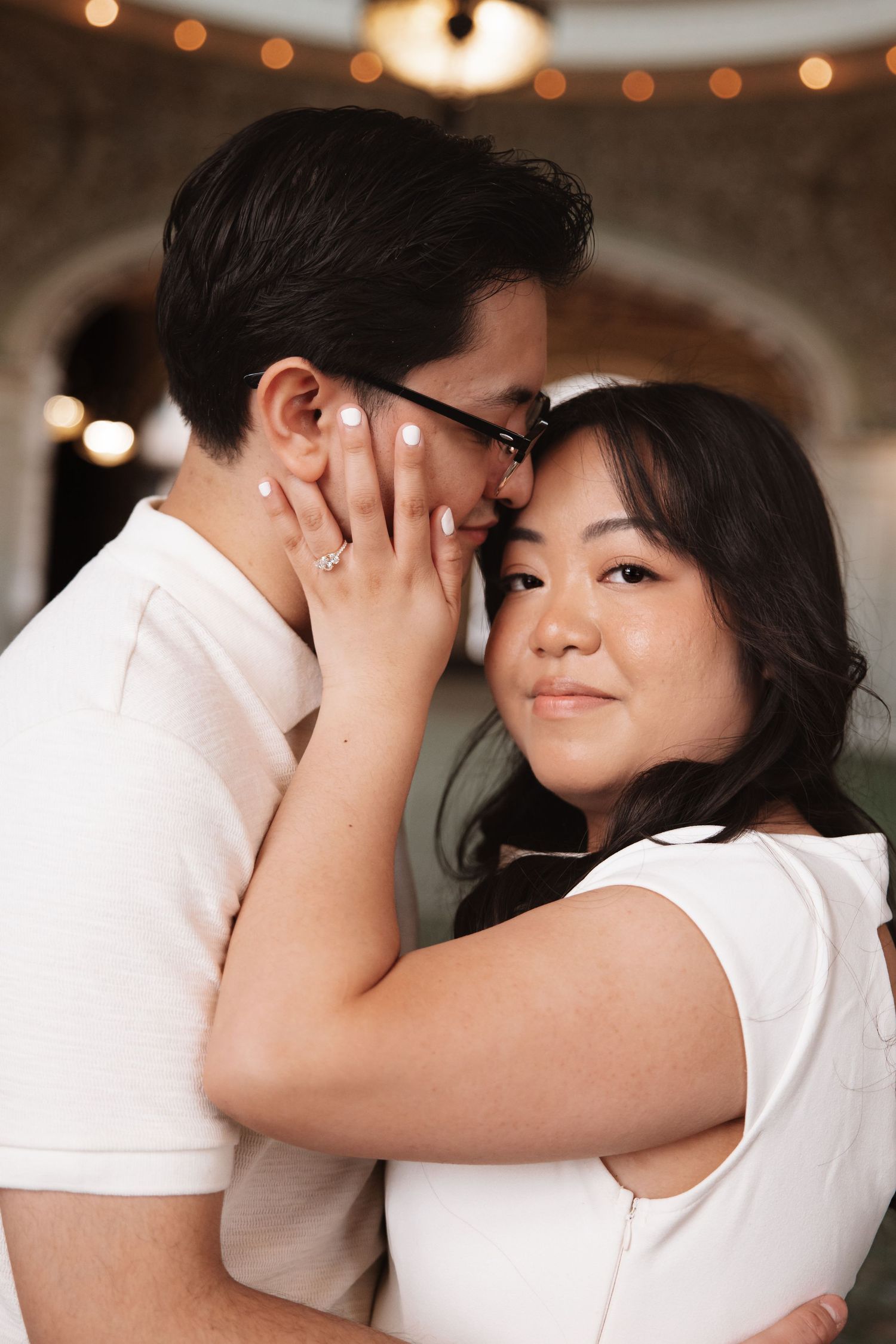 Couple sharing intimate moment, woman with engagement ring gently cupping man's face under warm bokeh lights.