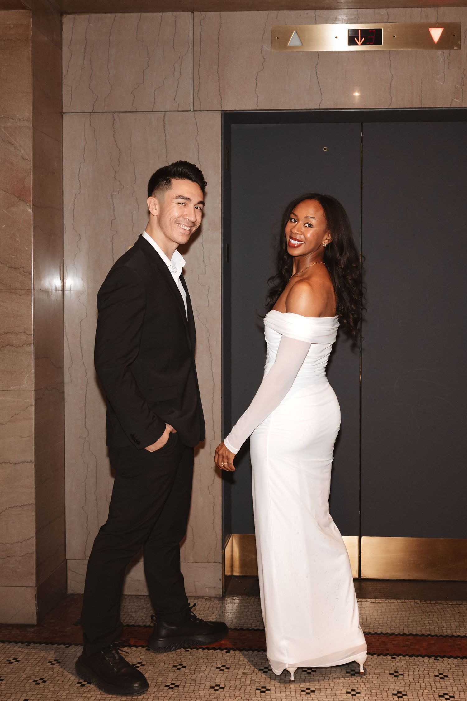 Stylish interracial couple holding hands and smiling outside a luxury hotel elevator, dressed in formal attire.Chicago Wedding Photographer