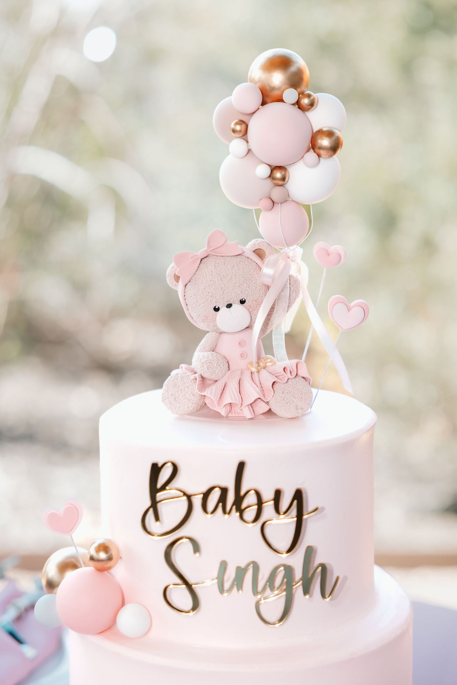 White Baby Singh cake with pink teddy bear ballerina topper holding gold and pink balloons.