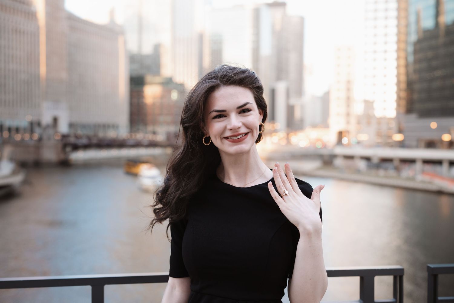 Smiling brunette woman in black dress showing engagement ring on Chicago Riverwalk at golden hour.