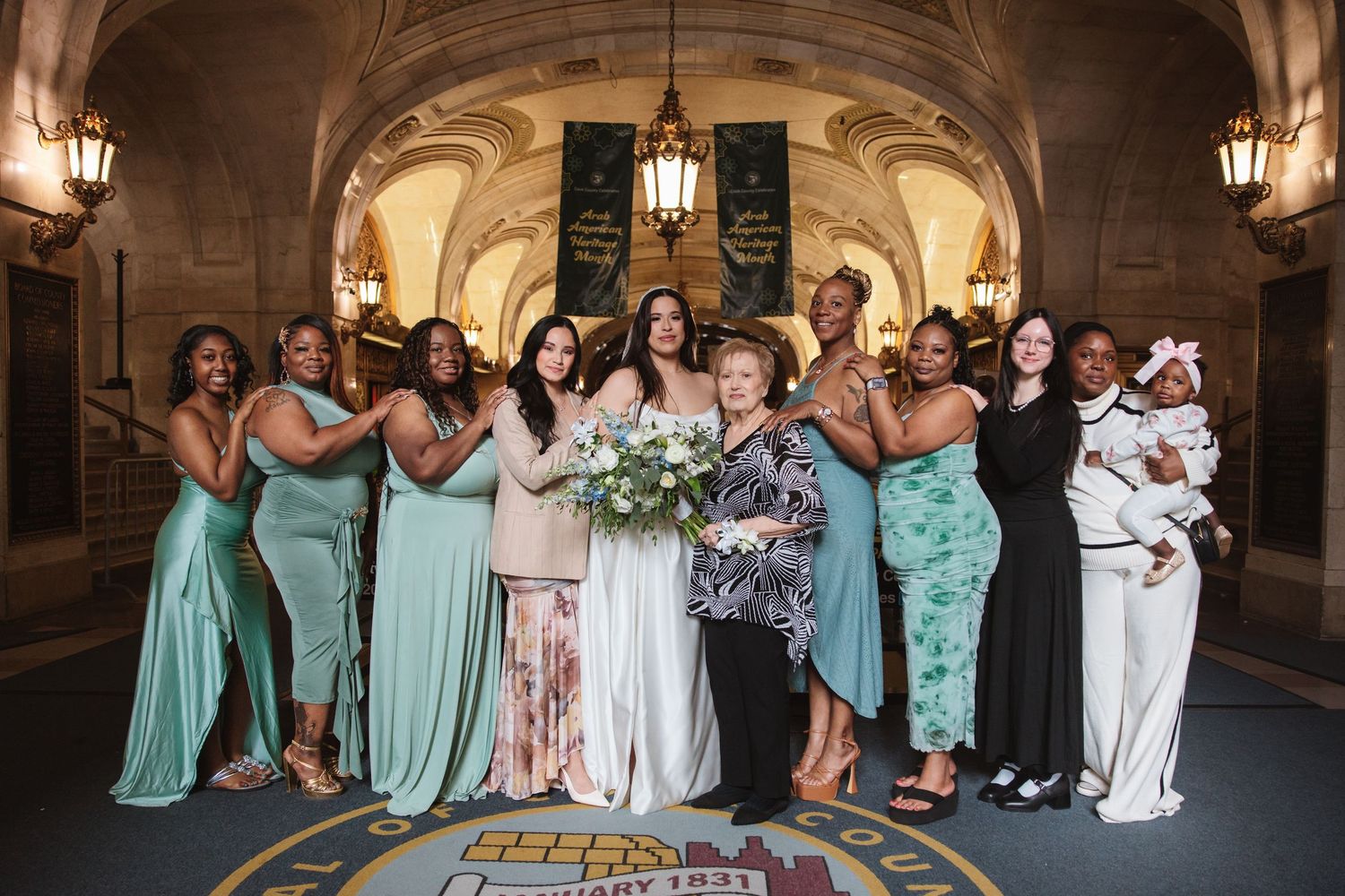 Bride poses with bridesmaids in teal dresses and guests in elegant wedding party photo inside ornate venue.