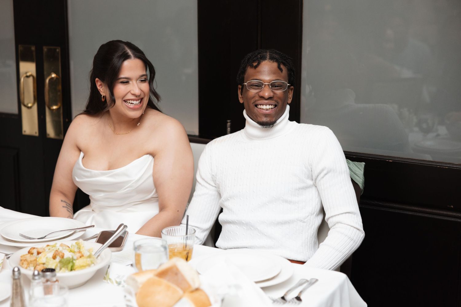 Smiling couple in white outfits seated at a wedding reception dinner table.