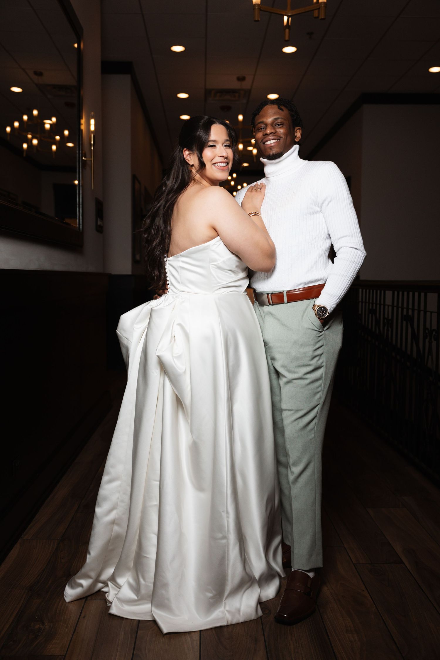 Smiling bride and groom in white turtleneck pose together in dimly lit reception venue.