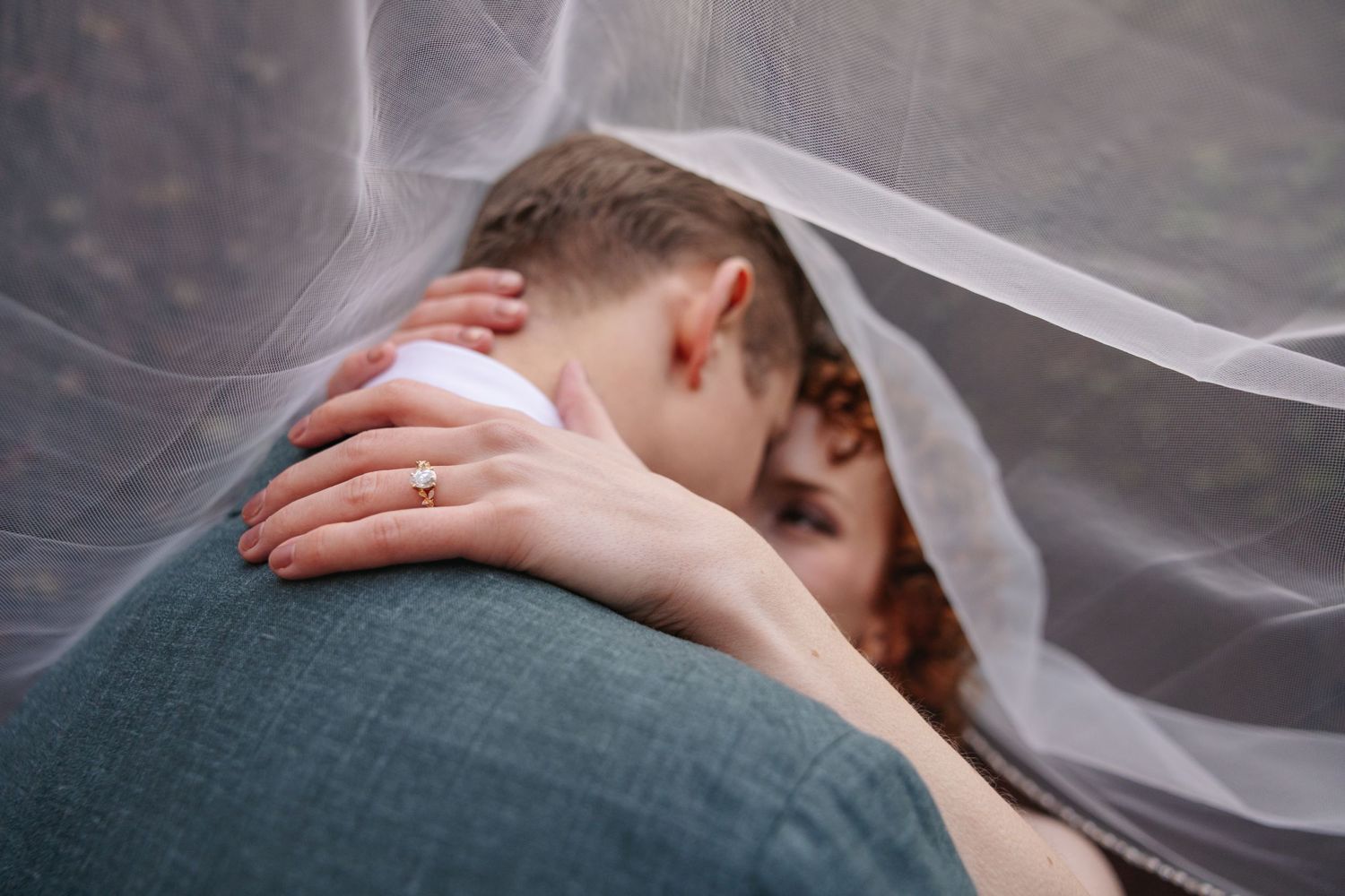 Close-up of bride and groom embracing under a sheer wedding veil, bride wearing a gold diamond ring.