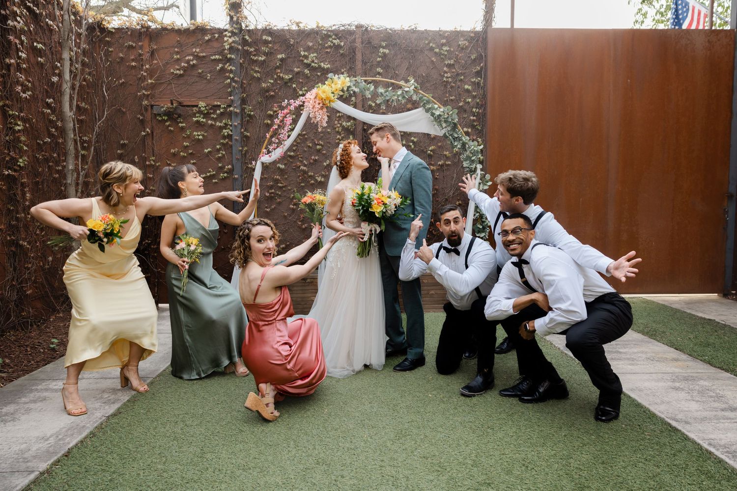 Wedding party strikes fun playful poses in front of floral arch at outdoor garden wedding celebration.