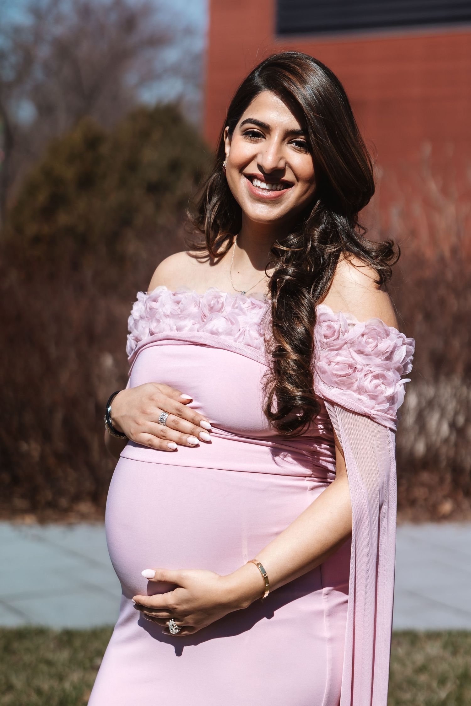 Glowing pregnant woman in pink floral off-shoulder dress smiles outdoors in bright sunlight.