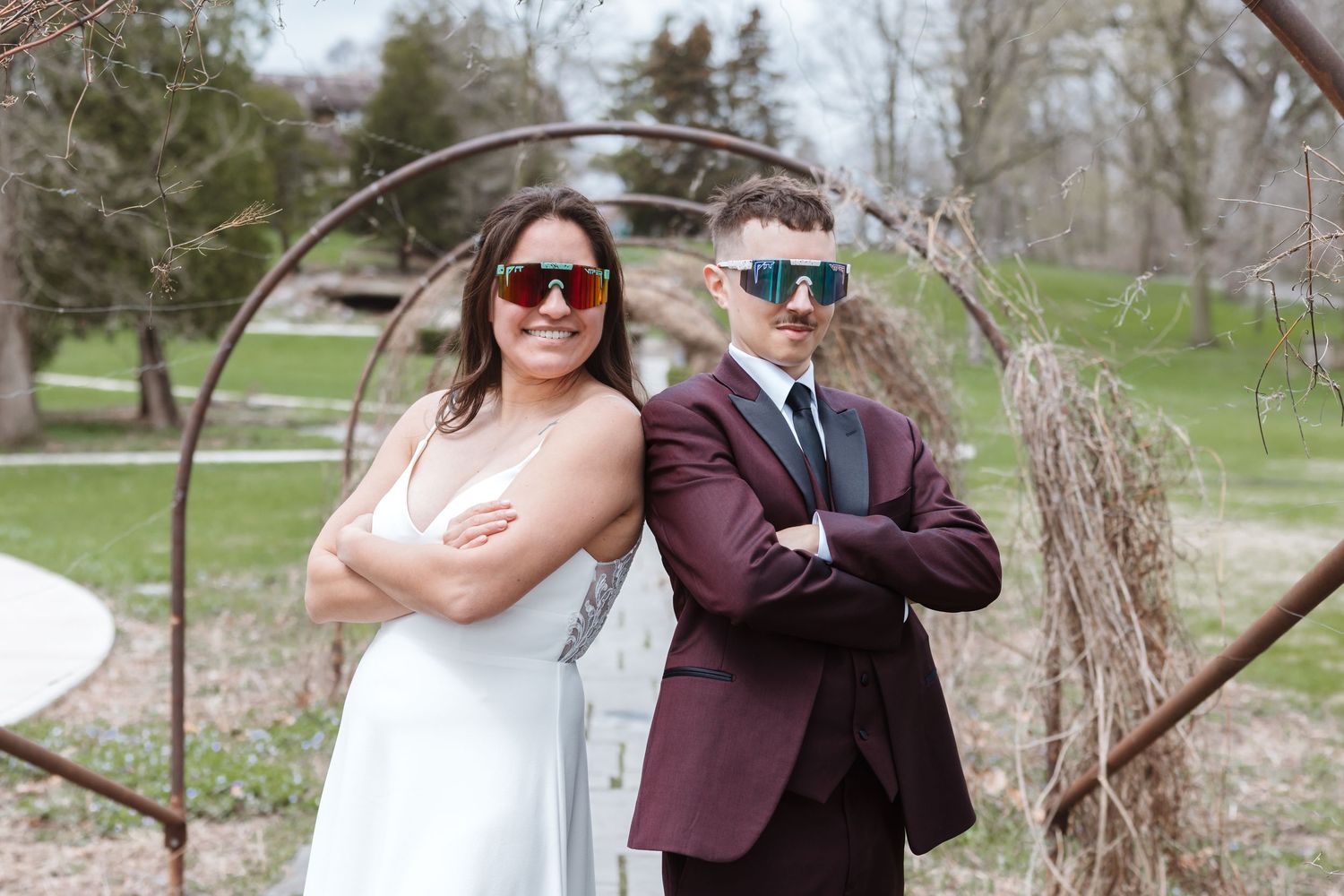 Bride and groom in wedding attire wearing colorful sunglasses, posing back-to-back with arms crossed outdoors.