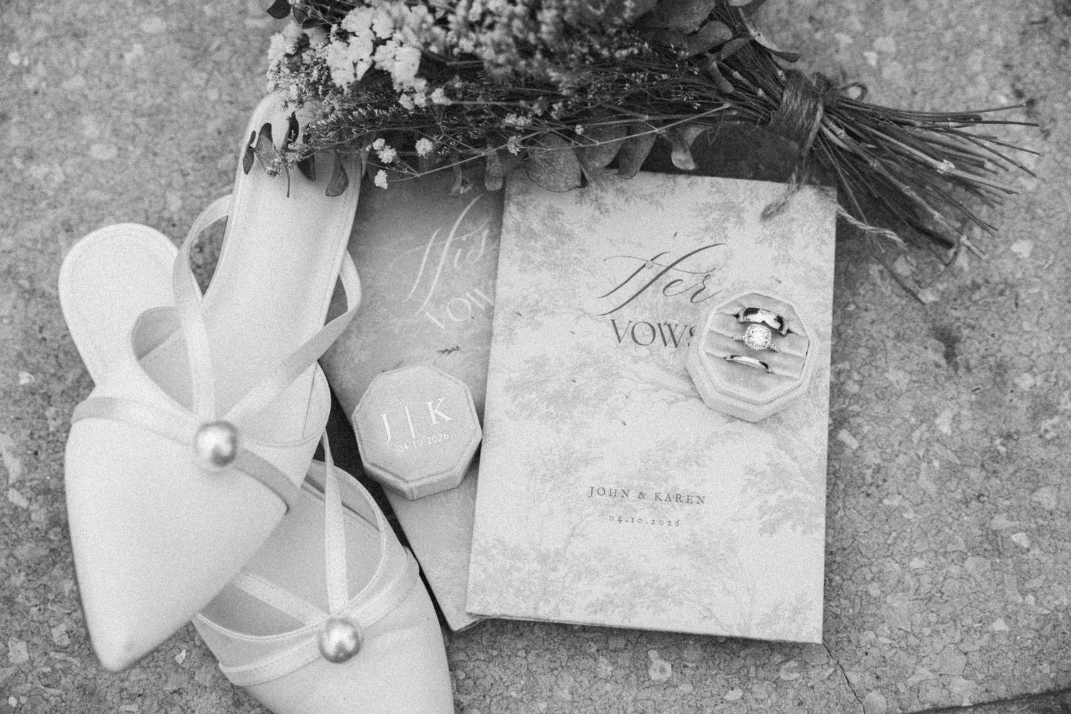 Black and white wedding flat lay with bridal heels, vow book, rings, and floral bouquet on stone surface.