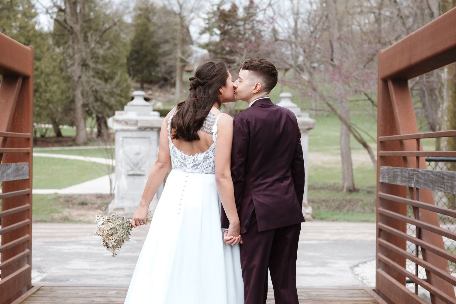 Couple kissing on wooden bridge in wedding attire, bride in white gown holding bouquet, groom in burgundy suit, park setting.