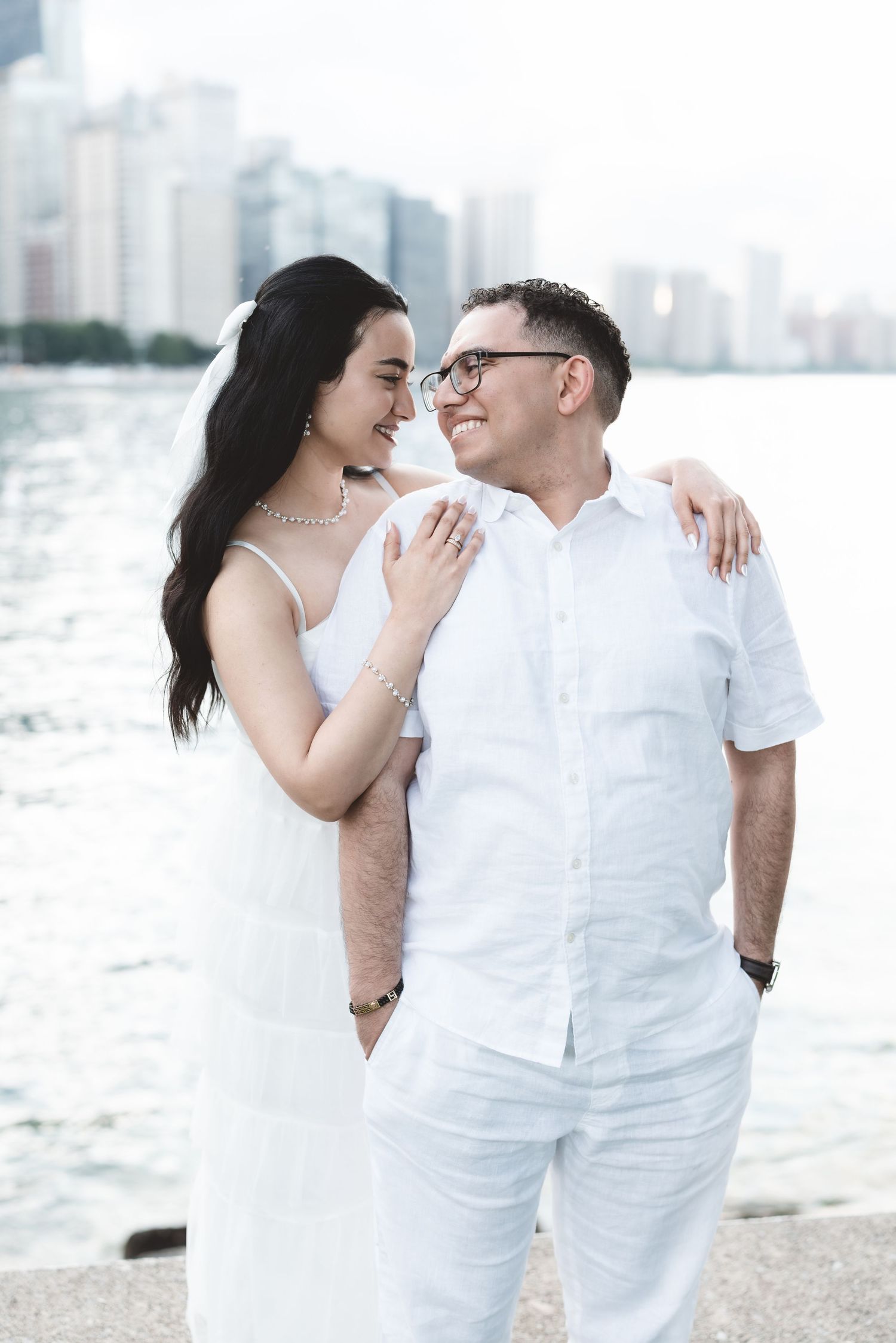 Couple in matching white outfits smiling at each other by a waterfront with city skyline in background.