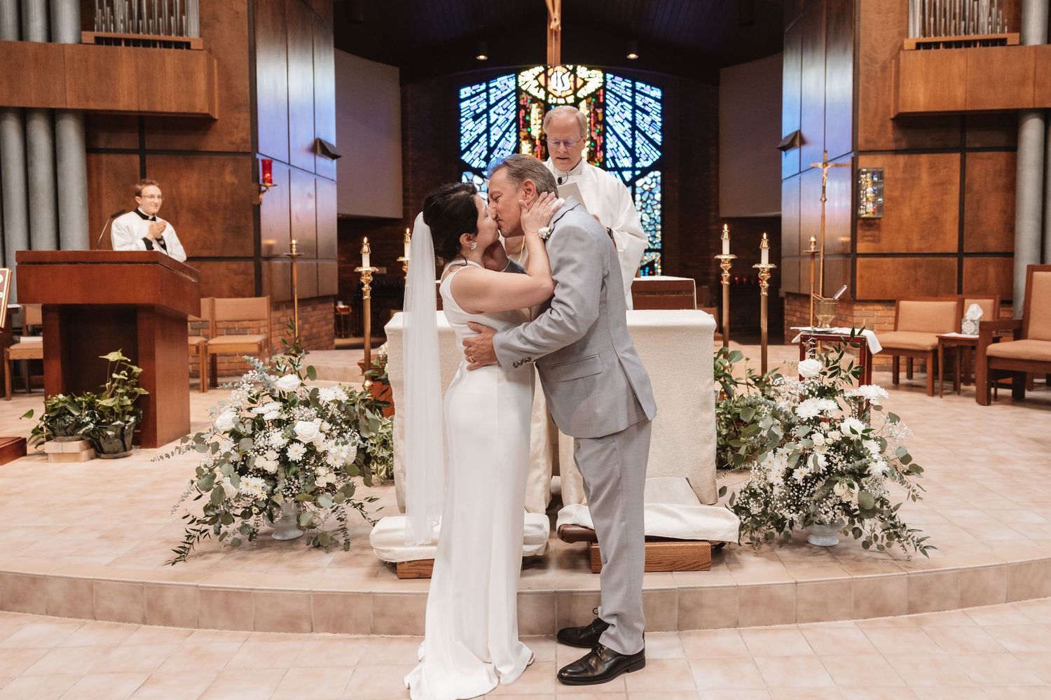 Newlyweds share first kiss at church altar surrounded by white floral arrangements during wedding ceremony.