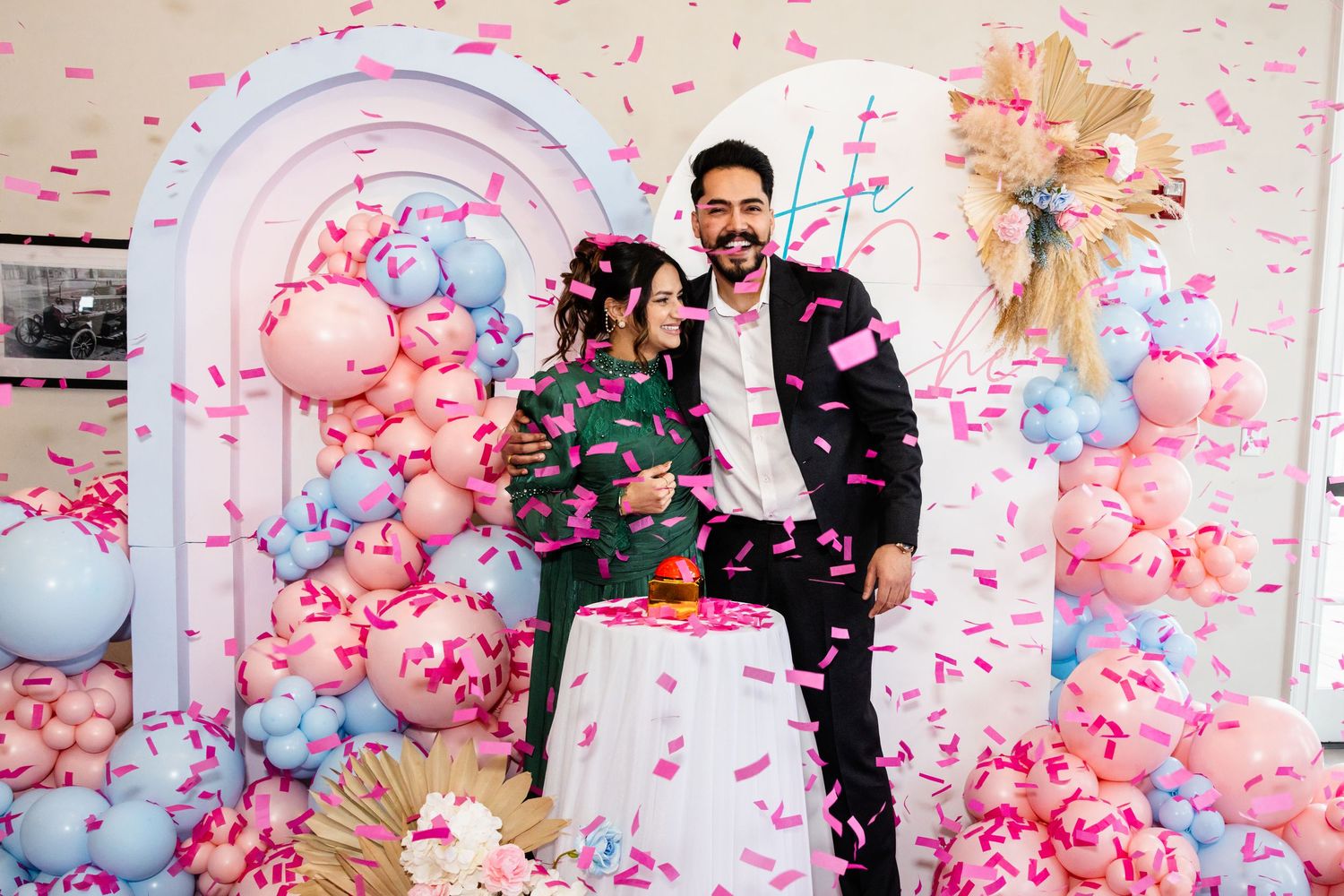 Couple celebrating gender reveal with pink confetti and pink and blue balloon decorations.