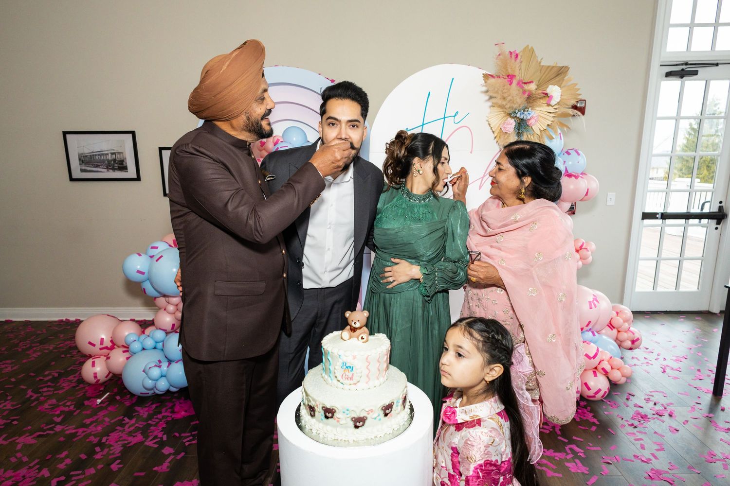 South Asian family celebrates gender reveal with cake cutting ceremony, surrounded by pink and blue balloon decorations.
