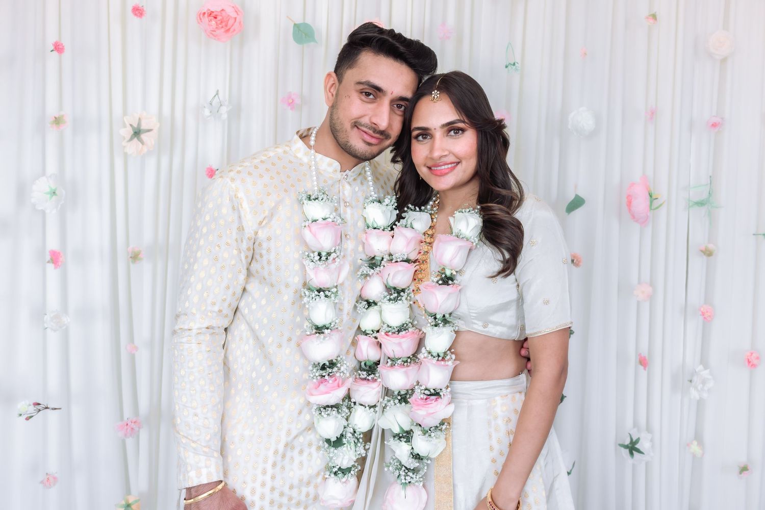 Indian bride and groom smile together wearing white outfits and floral garlands at their engagement.