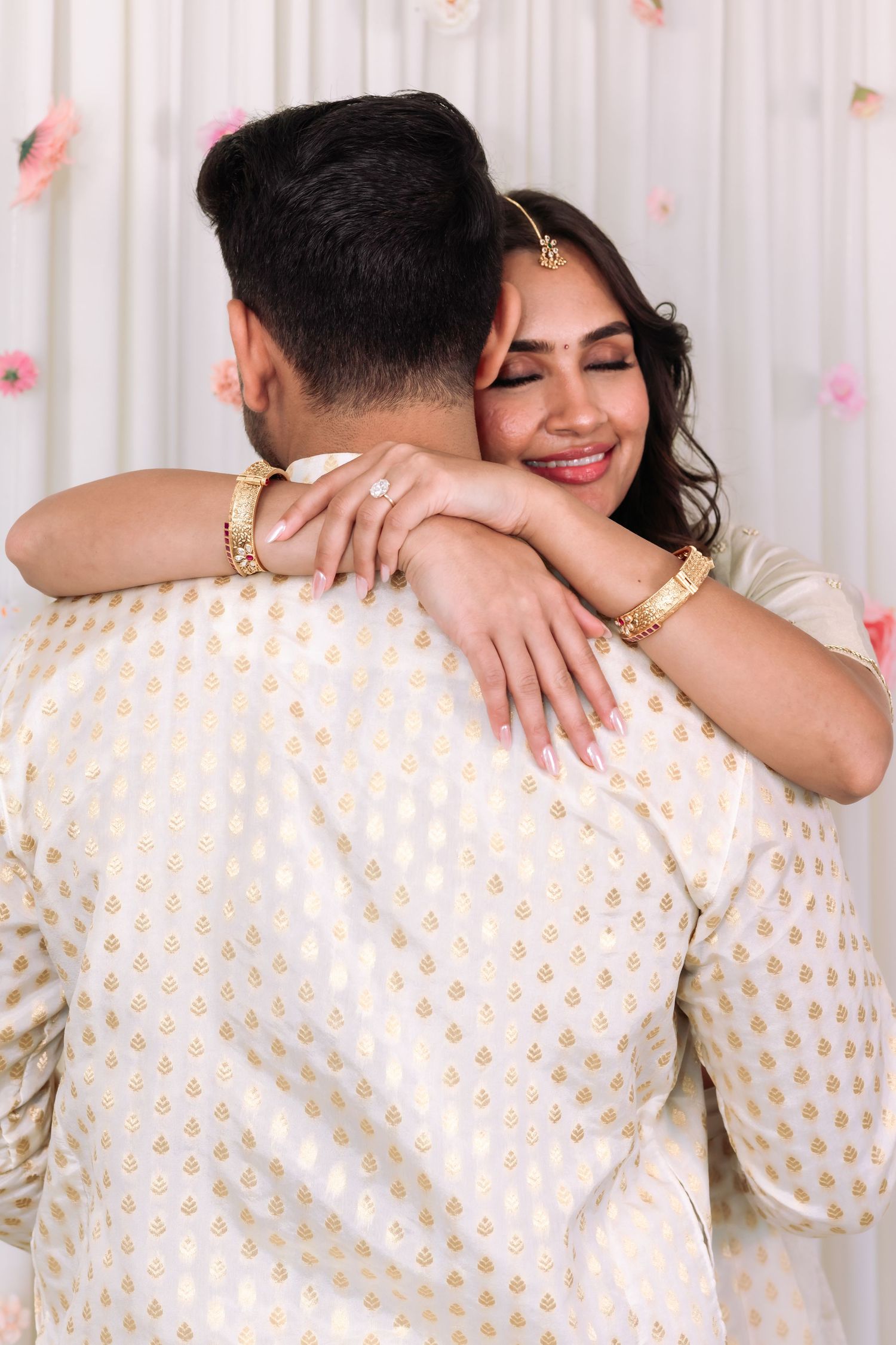 Bride in white lehenga hugging groom in gold kurta, showing engagement ring during Indian wedding ceremony.