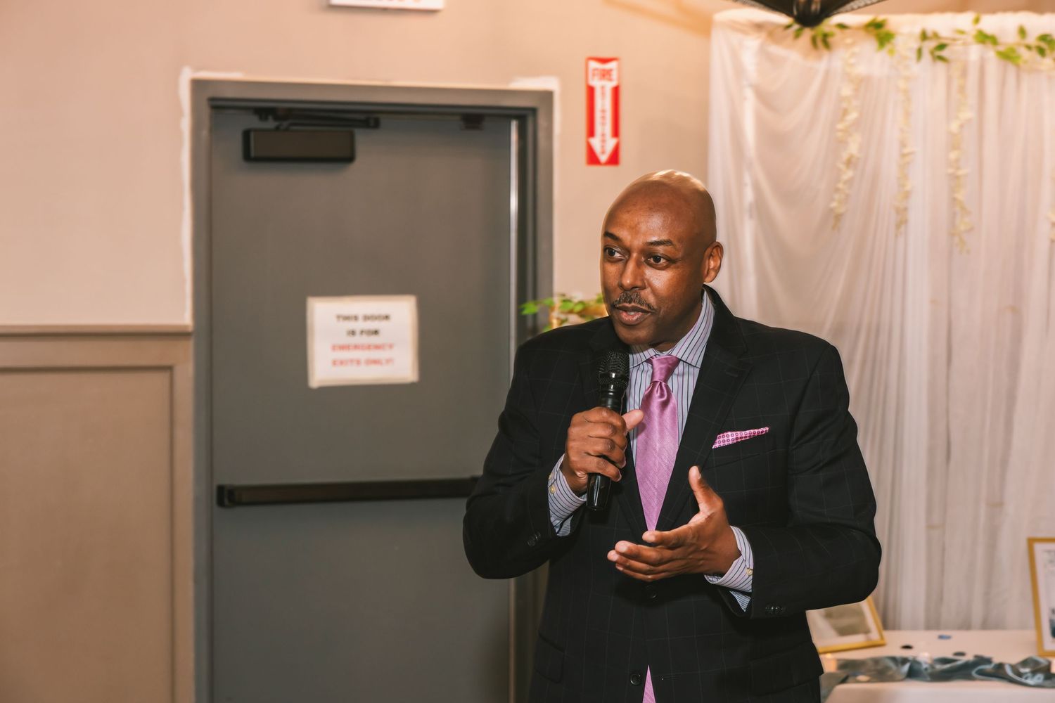 Well-dressed man in black suit and pink tie speaking into microphone at formal wedding reception.