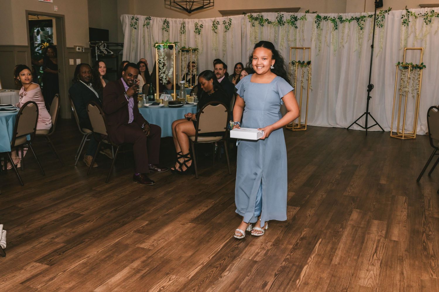 Smiling young girl in blue dress walks across wooden dance floor carrying a white gift box.