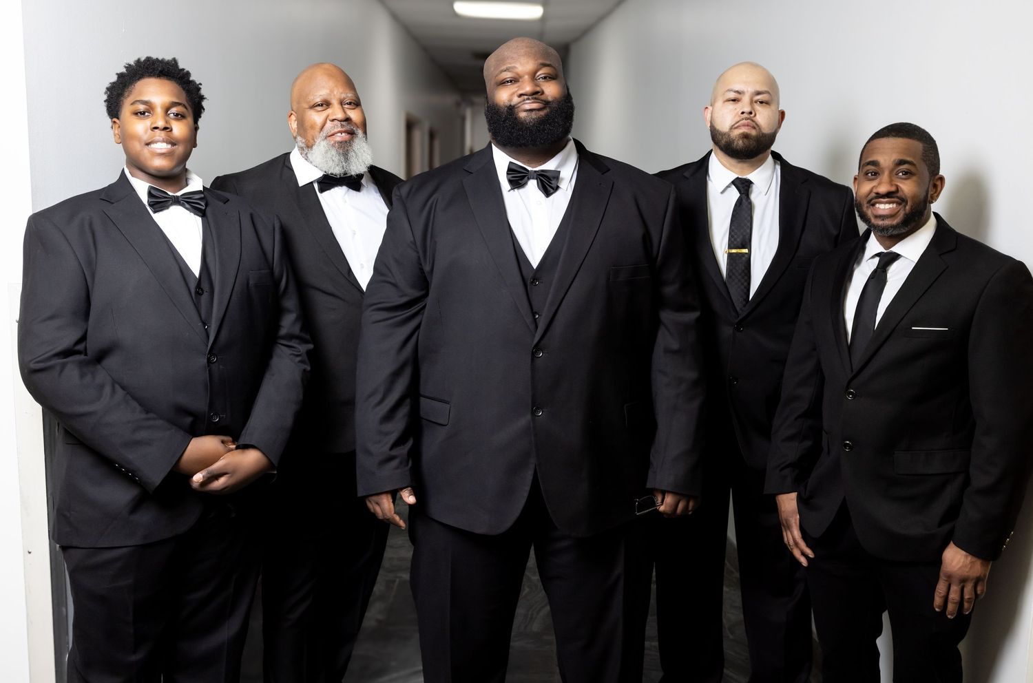 Five men in black tuxedos posing together as a groomsmen group in a bright corridor.