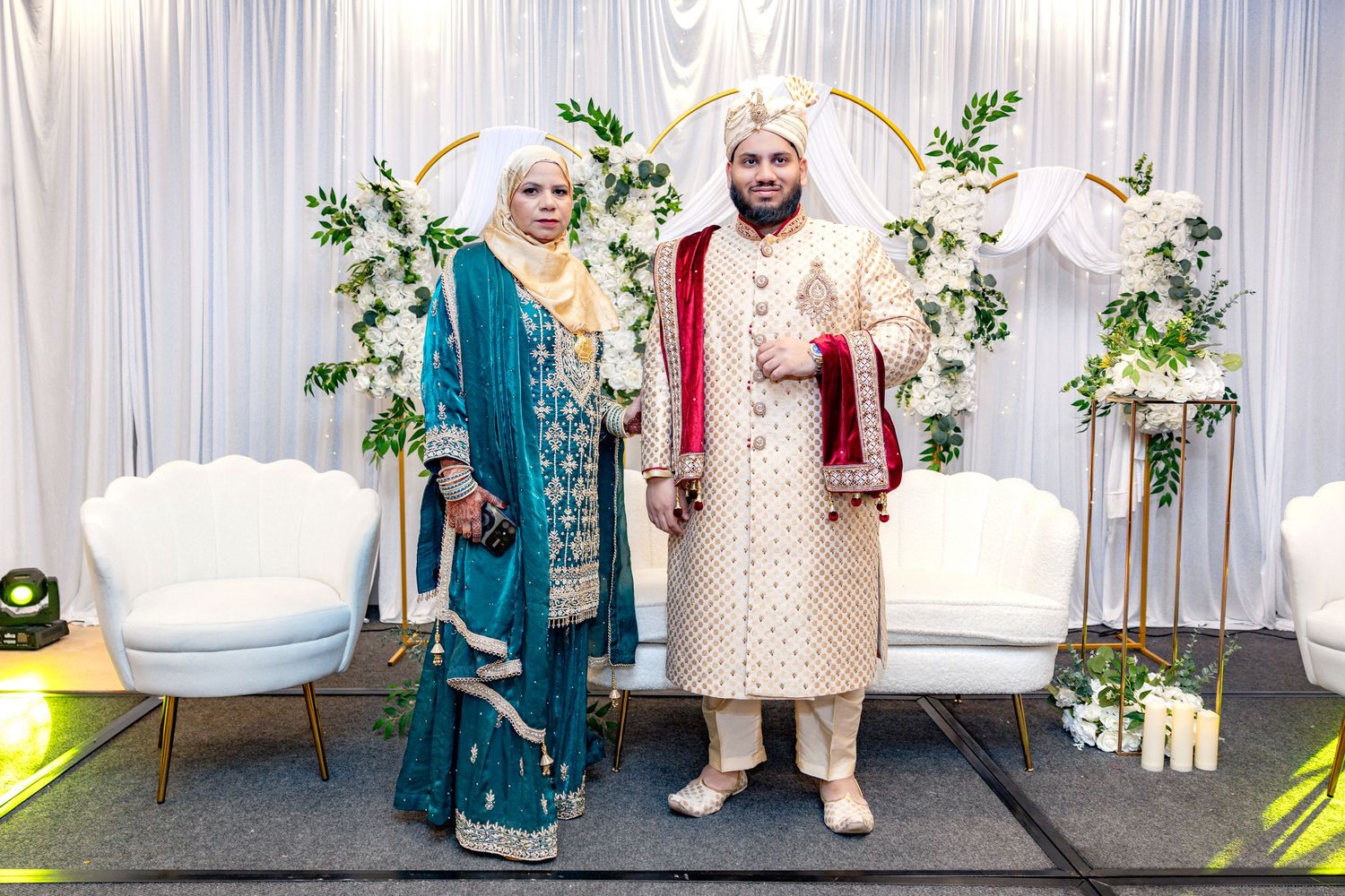 A couple in traditional South Asian wedding attire stands before an elegant floral arch at their ceremony.