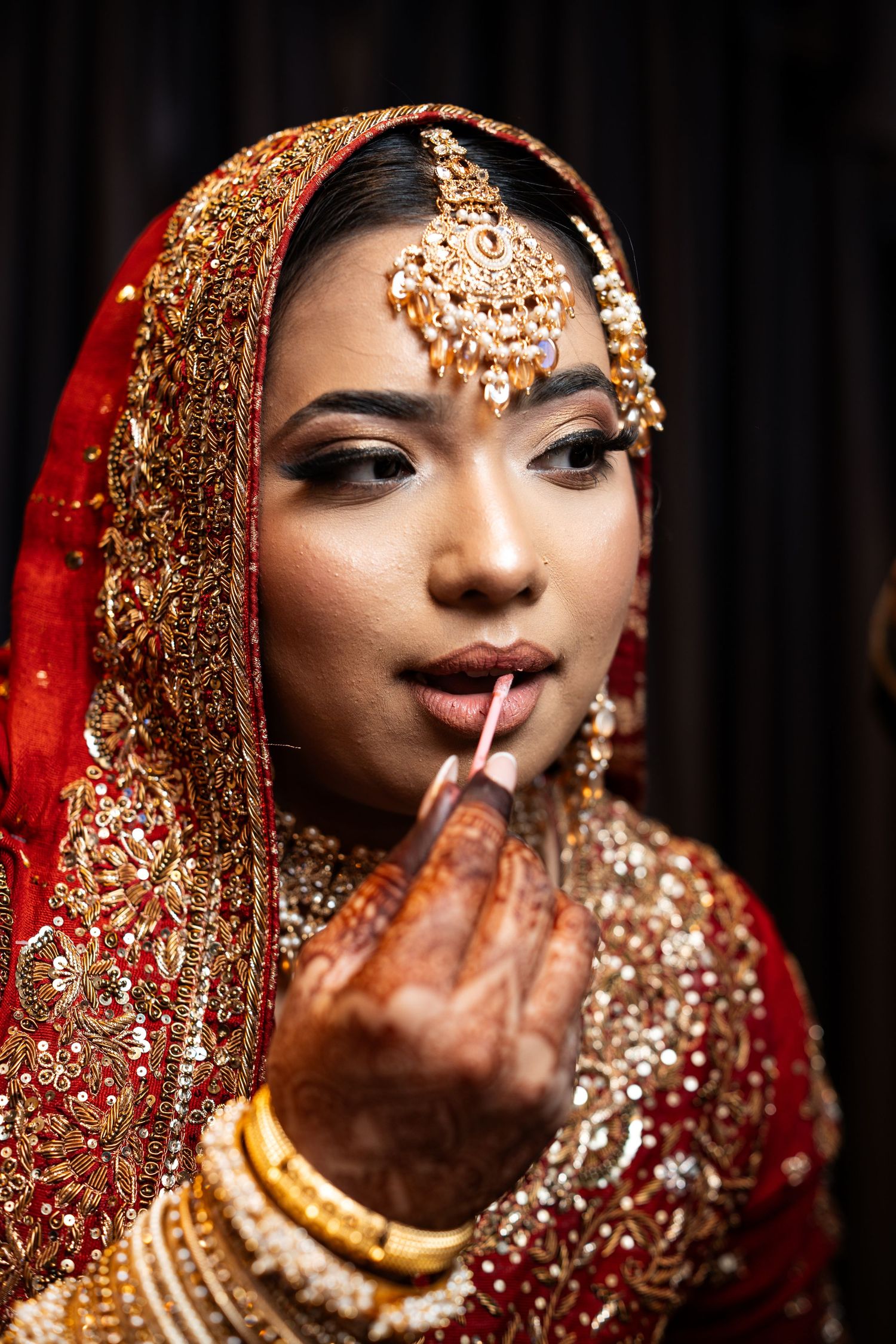 A stunning Indian bride in red and gold bridal attire gets her lipstick applied during wedding preparations.