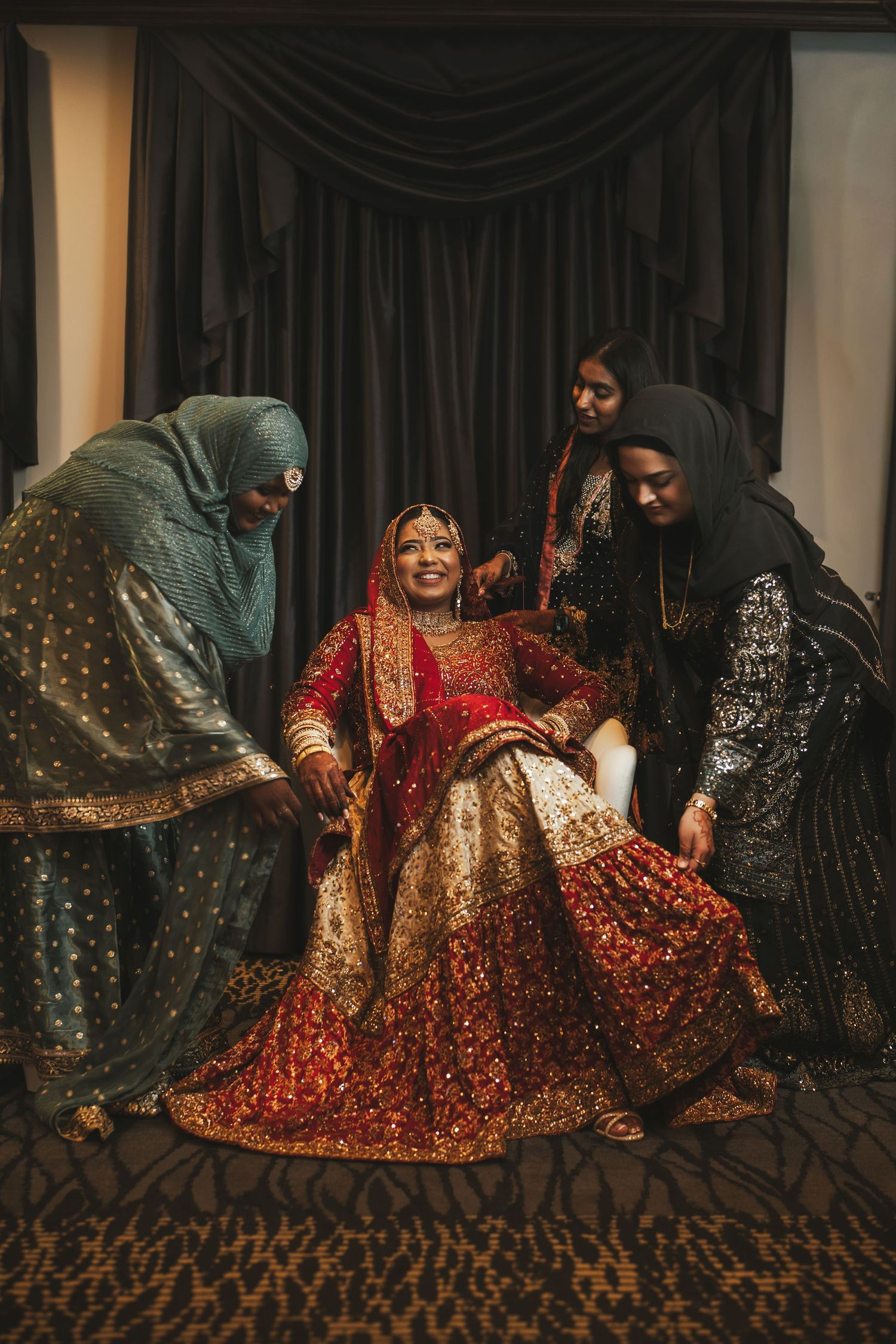 South Asian bride in red lehenga laughs joyfully as attendants help arrange her outfit.
