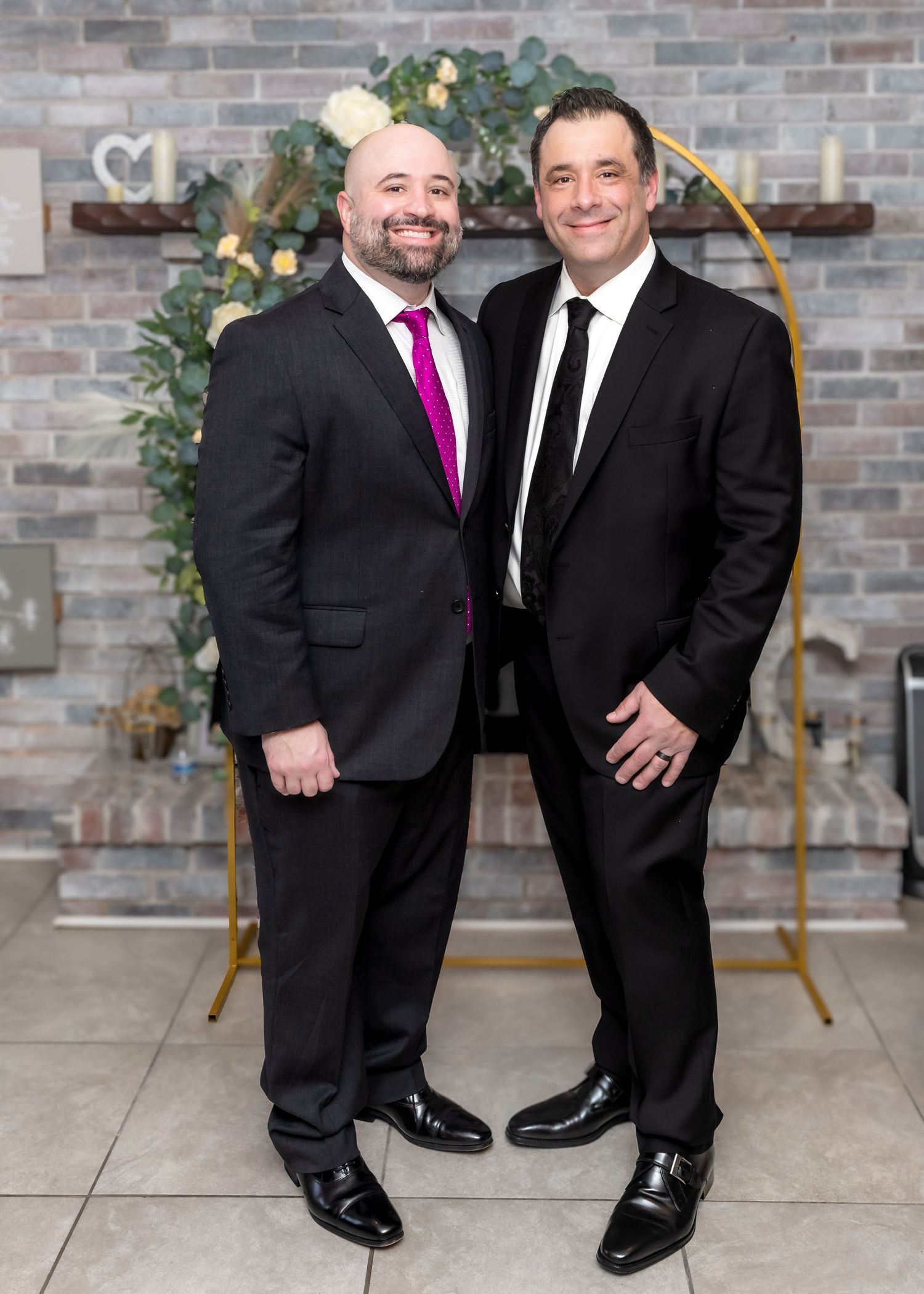 Two men in dark suits smiling together in front of a gold floral arch at a wedding.