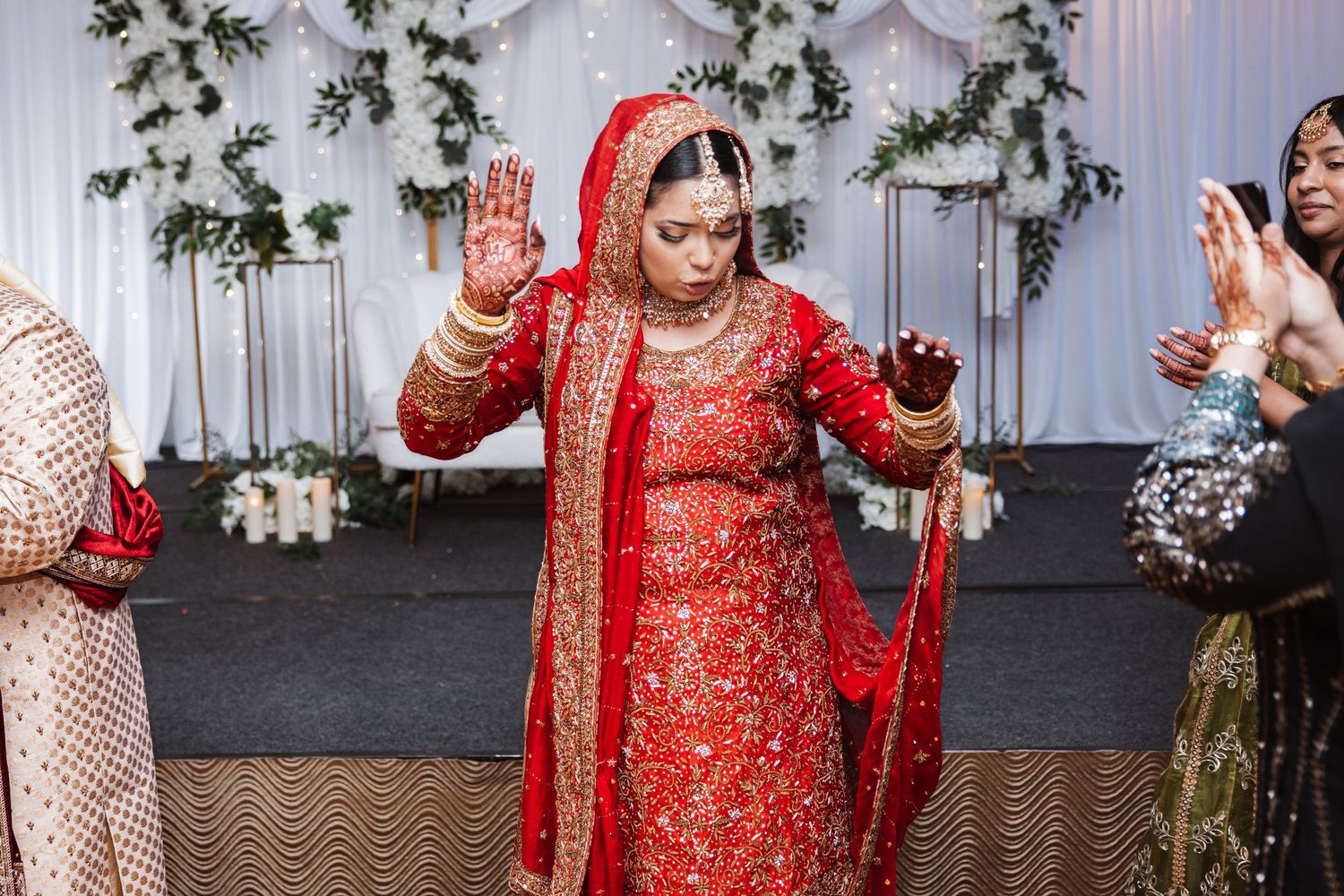 Bride in red embroidered lehenga with gold jewelry raises hands during South Asian wedding ceremony.