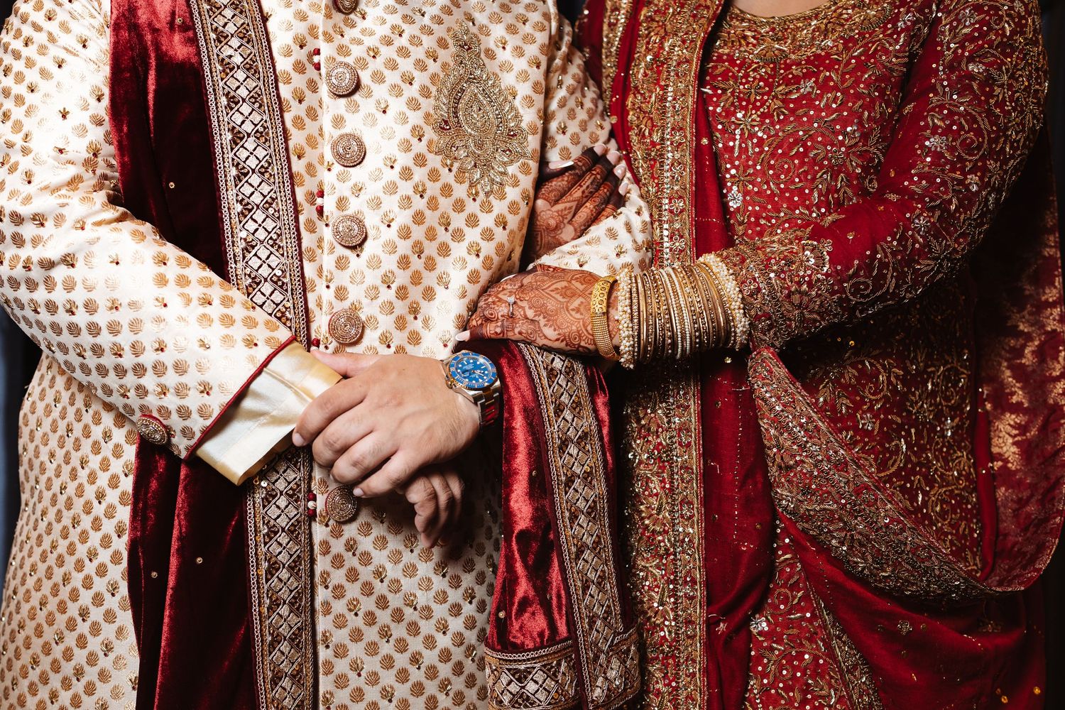 Close-up of South Asian bride and groom hands showing intricate mehndi, gold bangles, and blue watch.