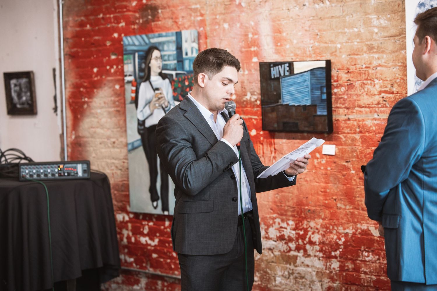 Man in suit speaks into microphone at indoor event with brick walls.