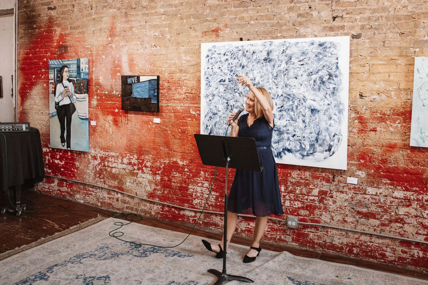Woman presenting at music stand in art gallery with large abstract paintings on exposed brick wall.