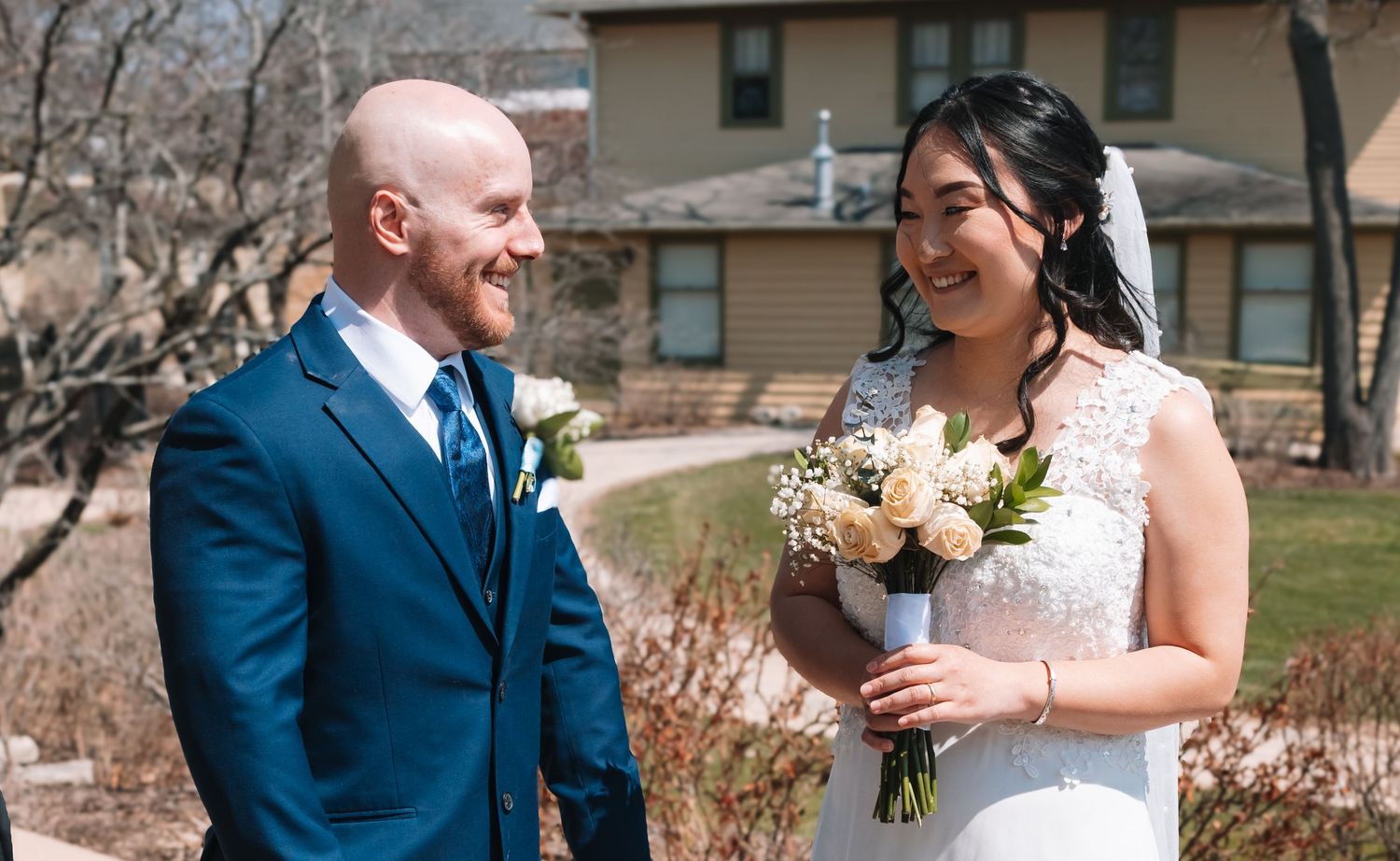 Smiling bride and groom share a joyful moment outdoors, bride holding a bouquet of white and cream roses.