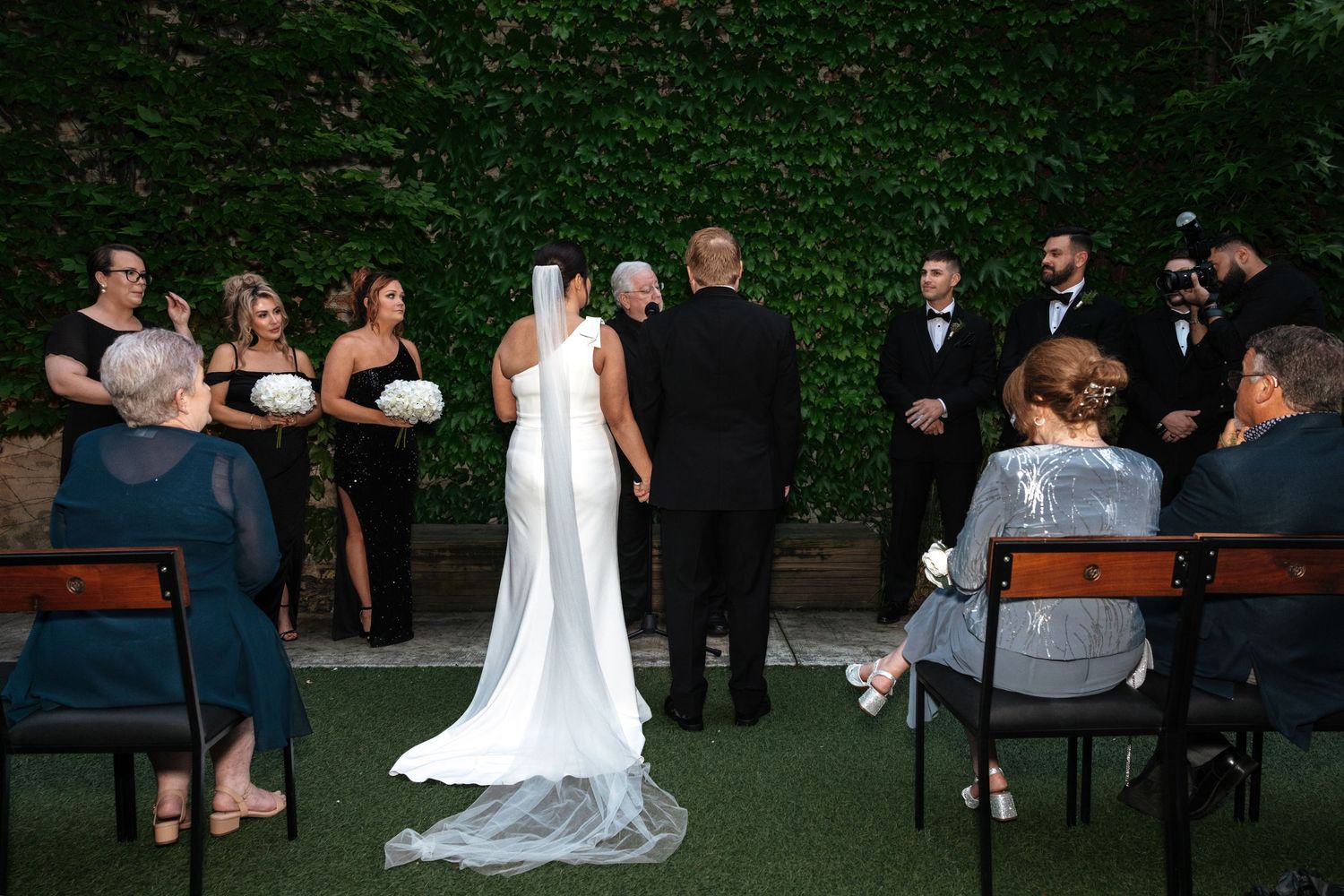 Wedding ceremony outdoors with bride, groom, officiant, bridesmaids, and groomsmen before ivy wall.