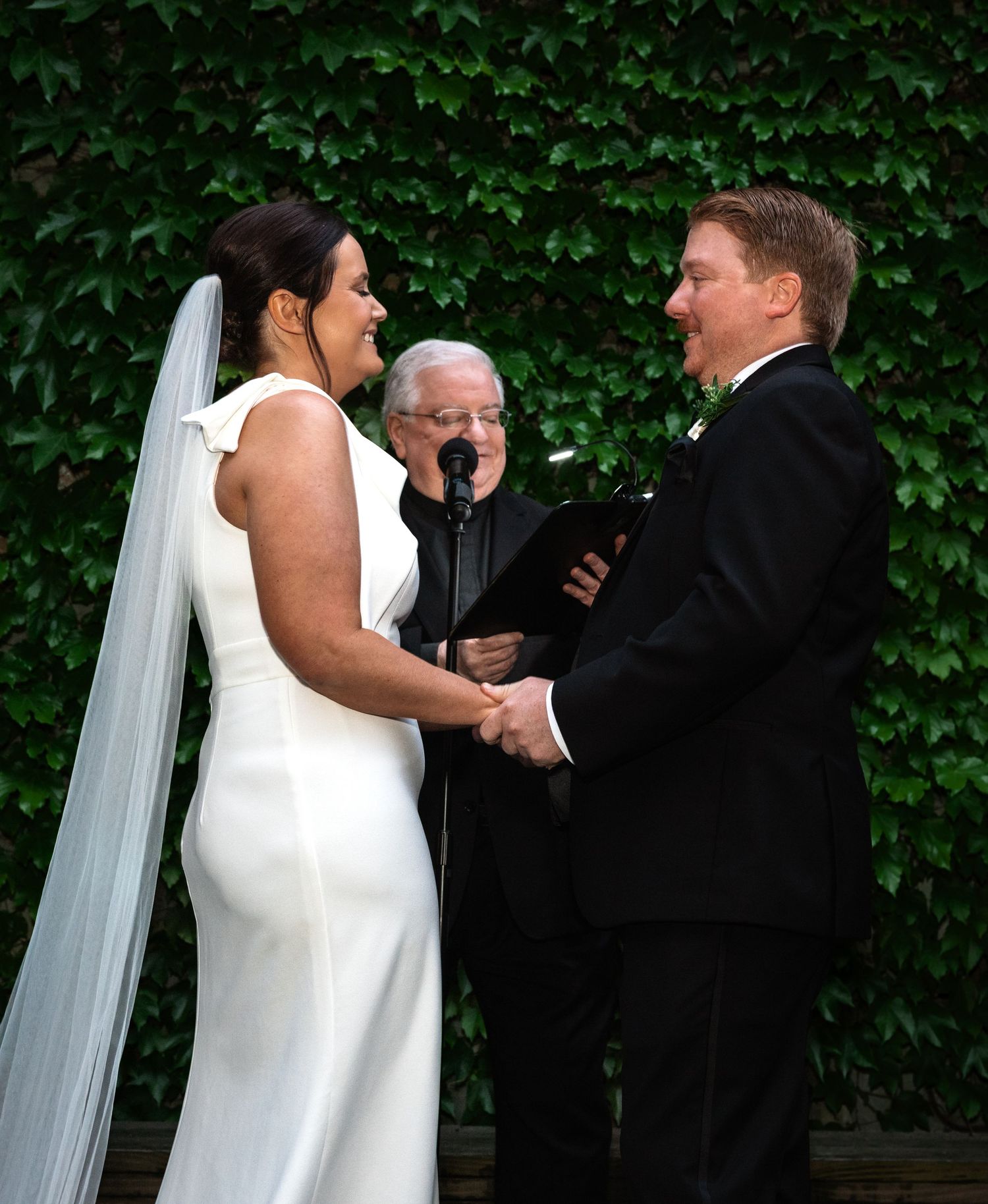 Smiling bride and groom hold hands before officiant during outdoor wedding ceremony with lush ivy backdrop.