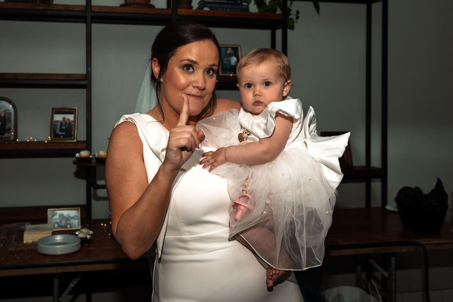 Bride in white dress holds baby girl in white outfit, posing with finger to lips in elegant indoor setting.