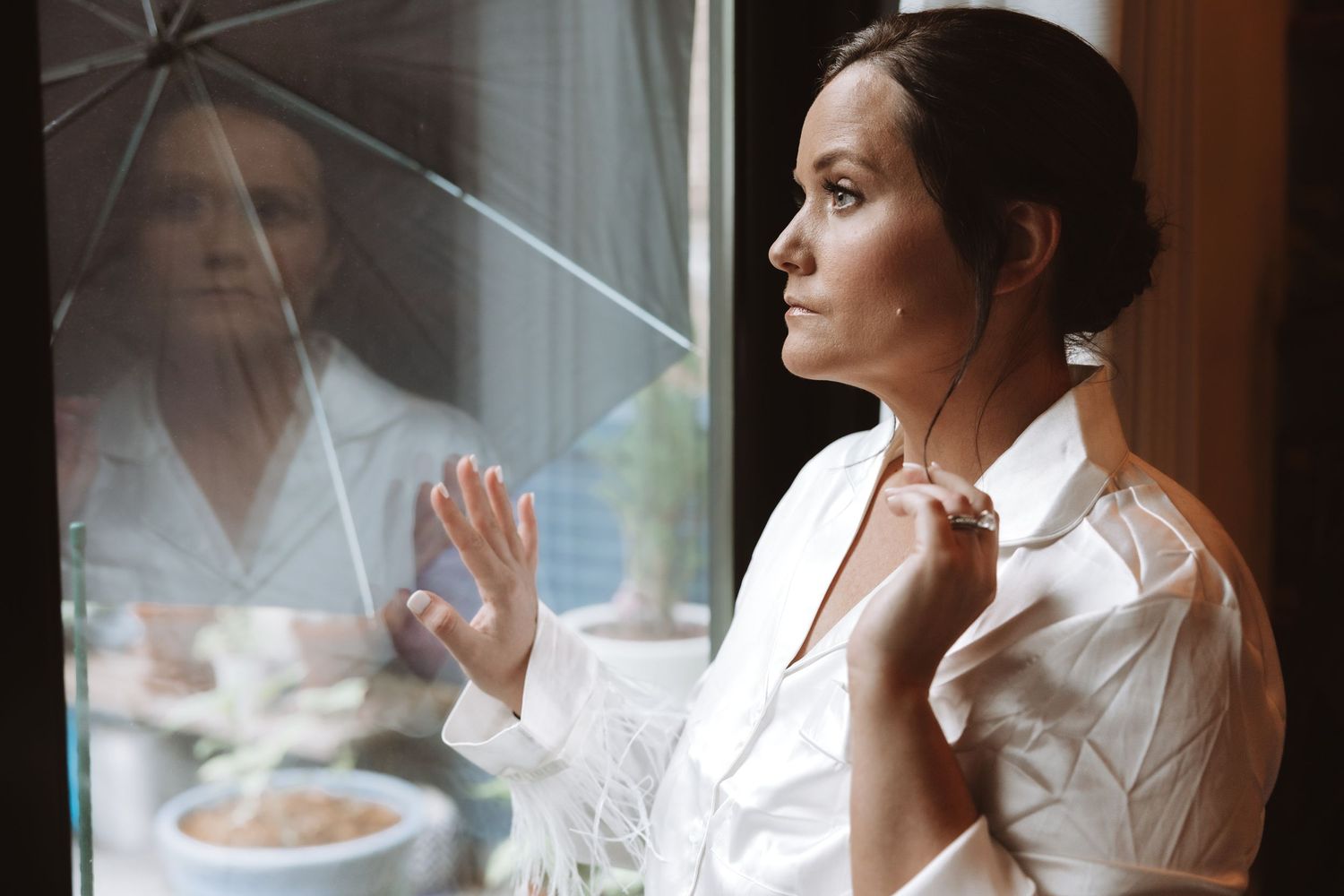Bride in white robe standing by window holding umbrella, looking thoughtfully outside.
