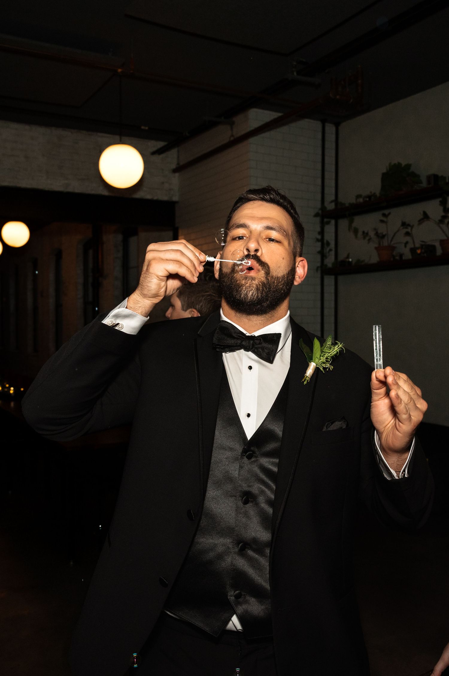 Dapper groom in black tuxedo blowing bubbles at indoor wedding reception with industrial-style decor.