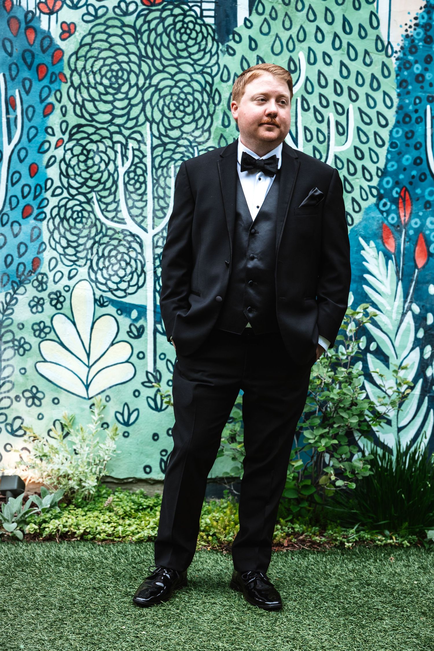 Groom in classic black tuxedo stands confidently before a vibrant painted garden mural backdrop.