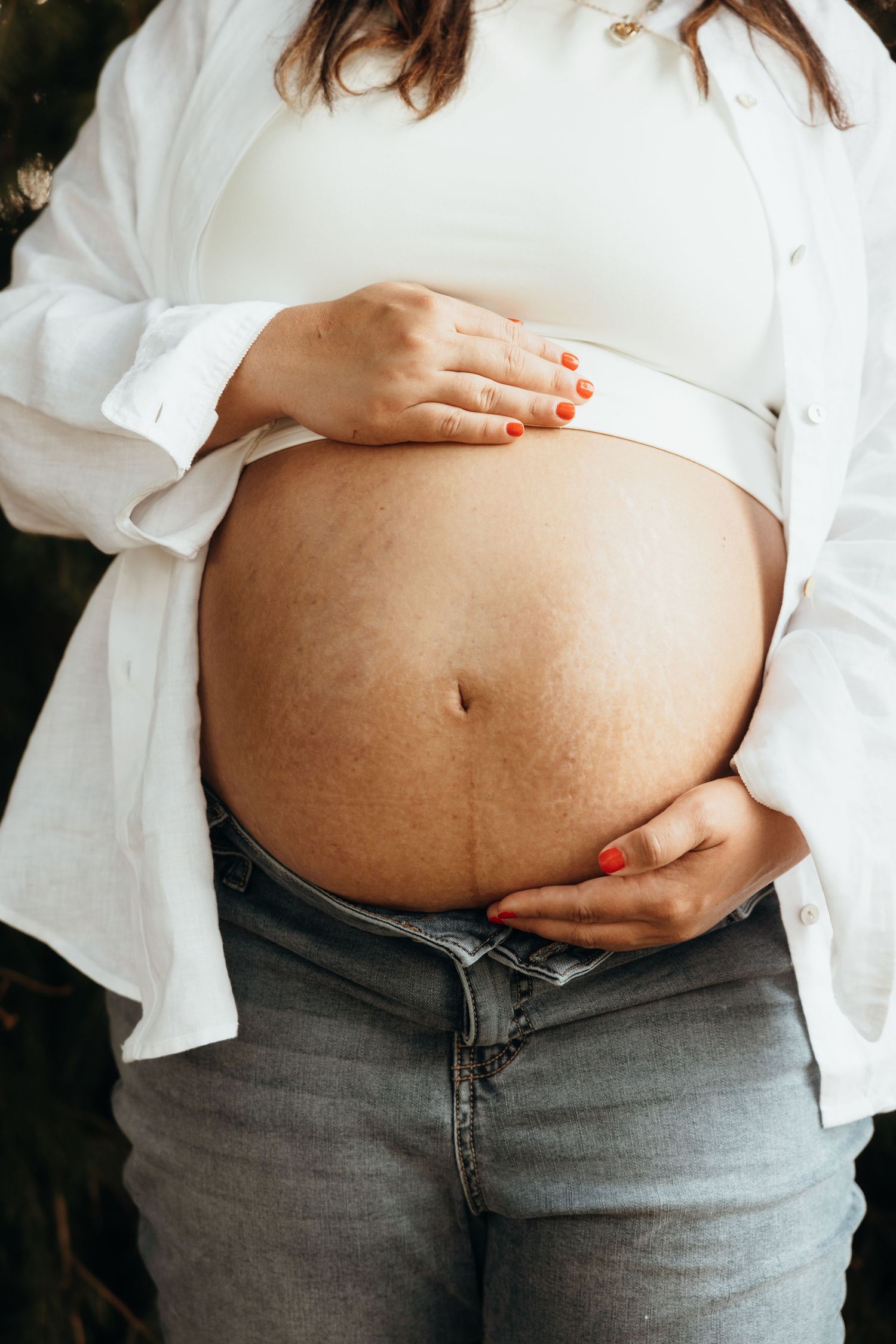 Close-up of pregnant woman in white shirt and jeans gently holding bare baby bump with red nails.