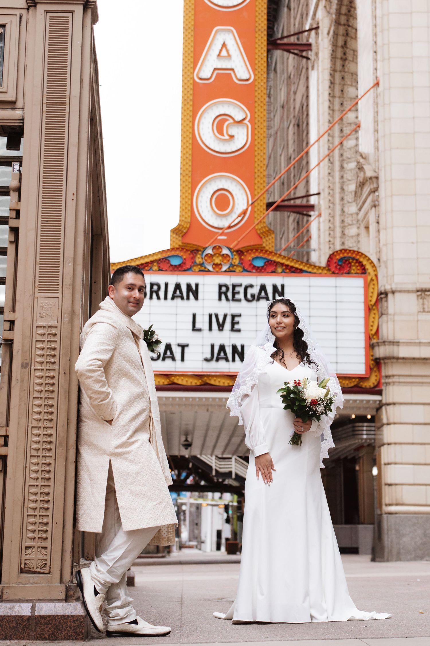 Bride in white gown and groom in cream sherwani pose by Chicago Theatre marquee sign.