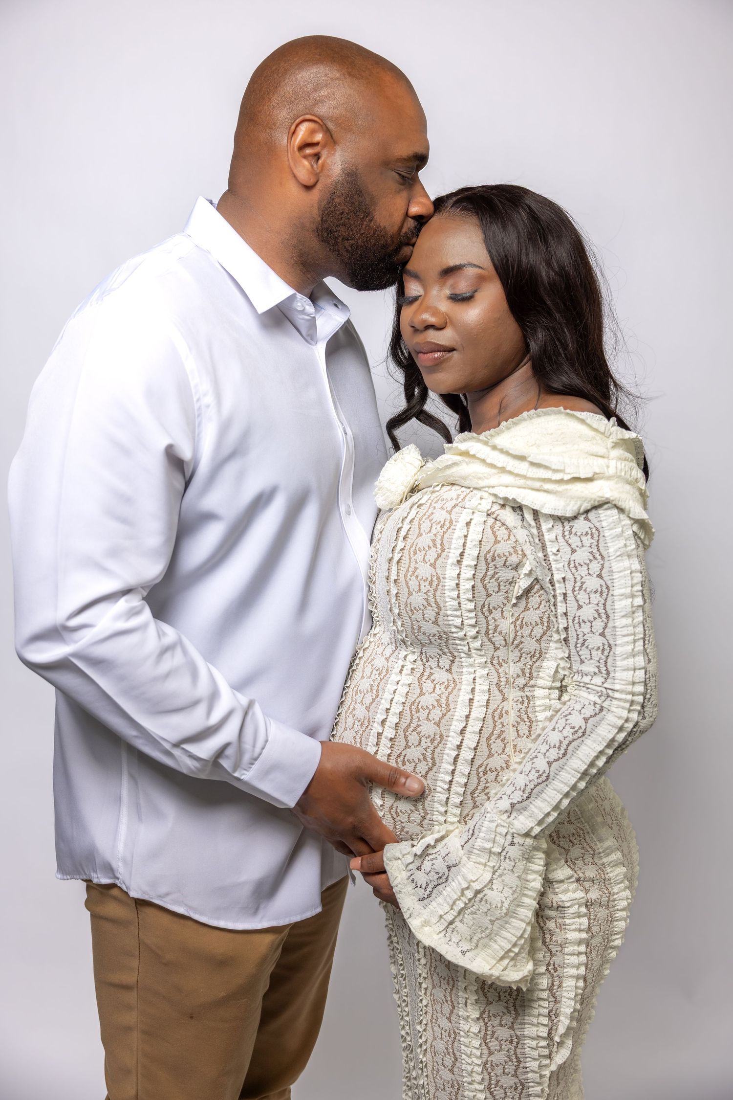 Man gently kissing pregnant partners forehead during intimate maternity photo session on grey background.