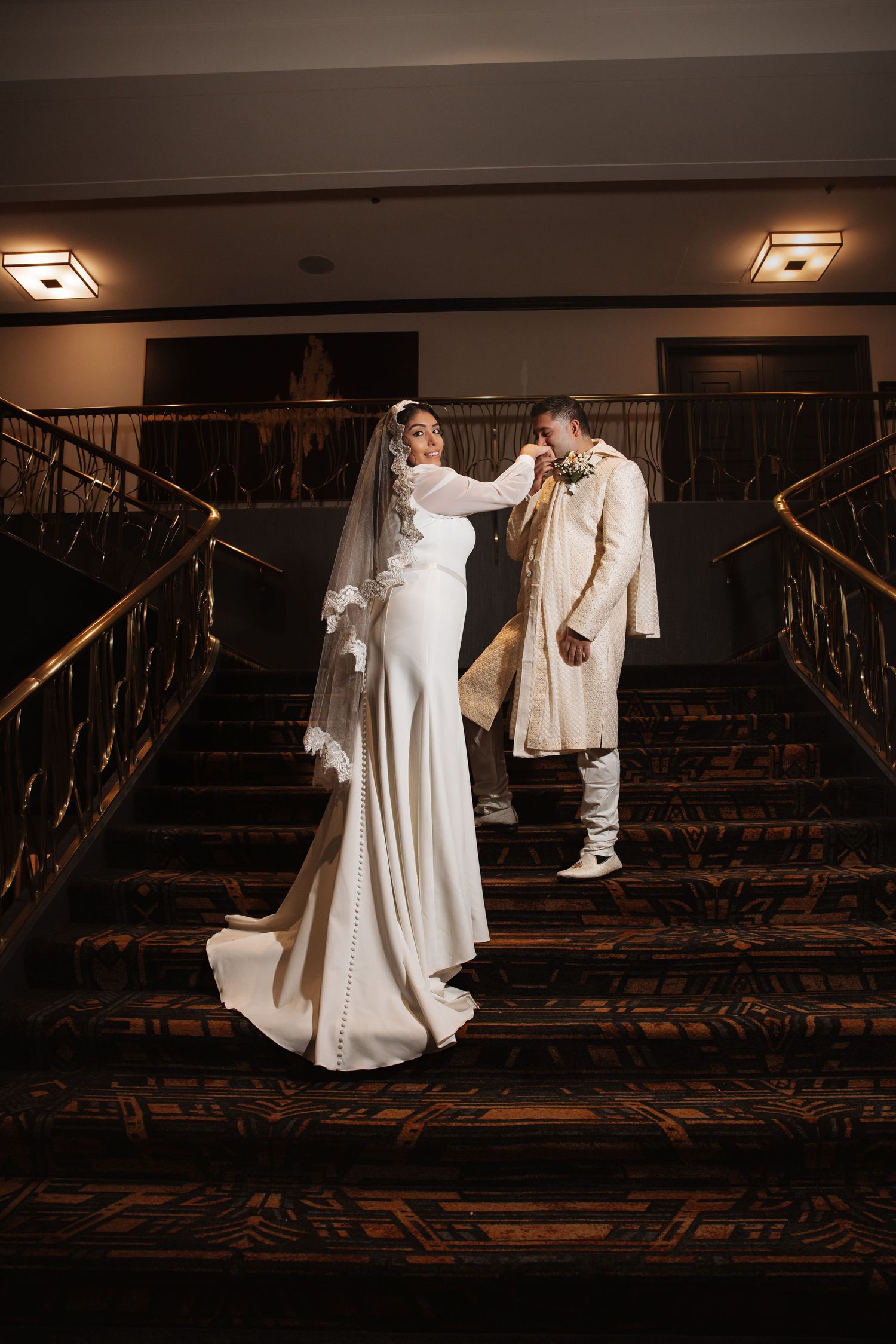 Bride in white gown adjusts grooms boutonniere on ornate carpeted staircase.