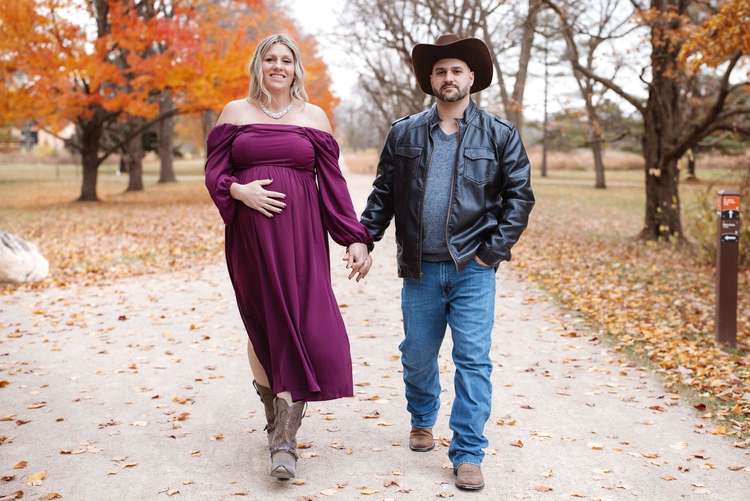 Expecting couple strolling hand-in-hand on autumn path, woman in burgundy maternity dress.