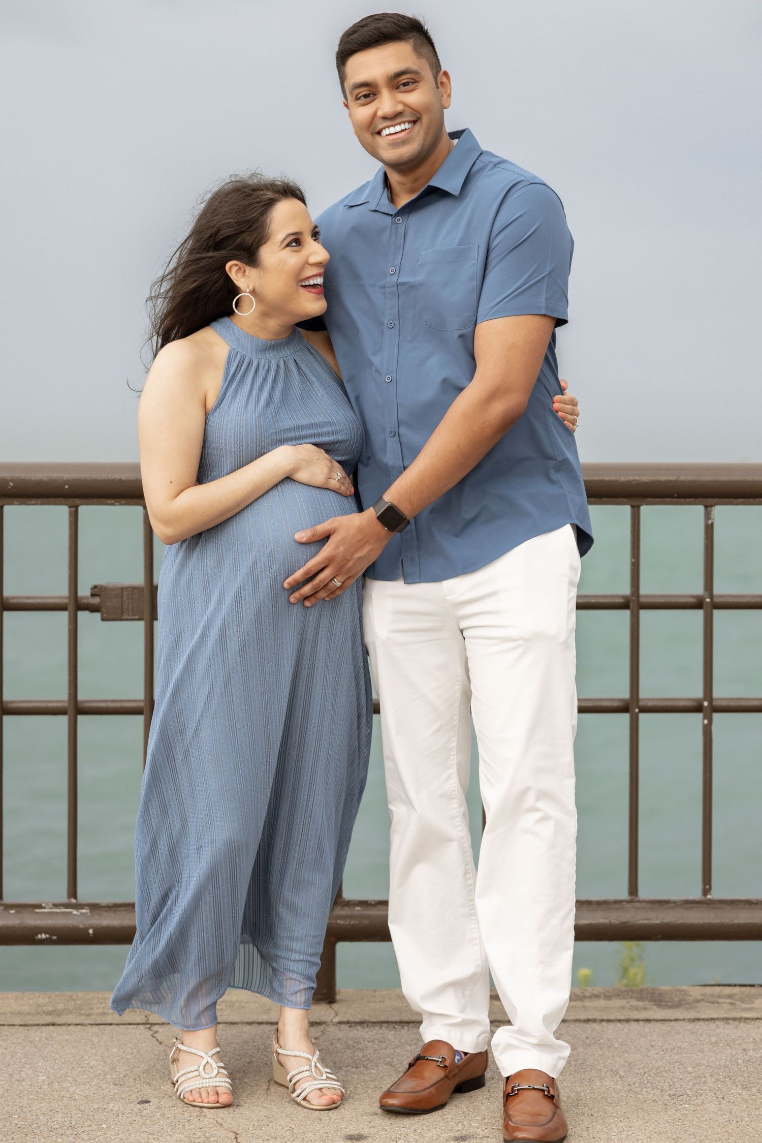 Happy pregnant couple embracing by metal railing with ocean in background.