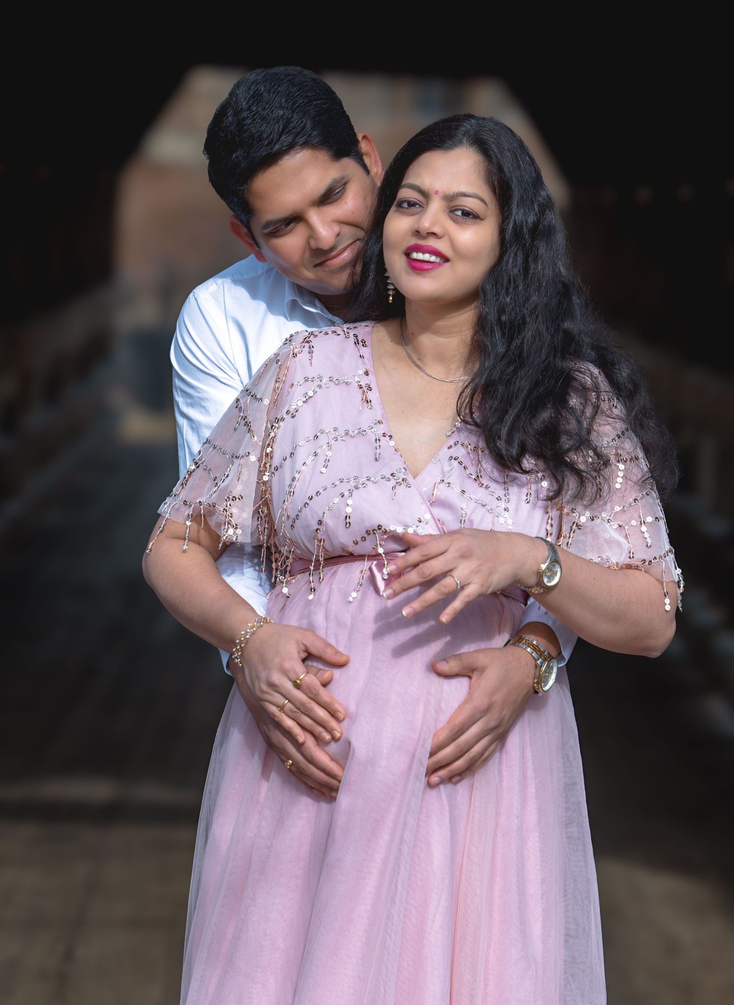 Couple posing for maternity photo shoot, man embracing pregnant woman in pink sequin dress, romantic outdoor portrait.