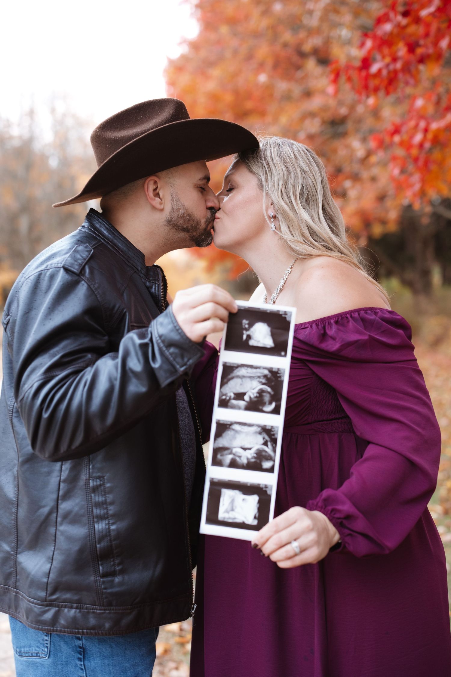 Expecting couple kisses while holding ultrasound photos surrounded by colorful autumn leaves.