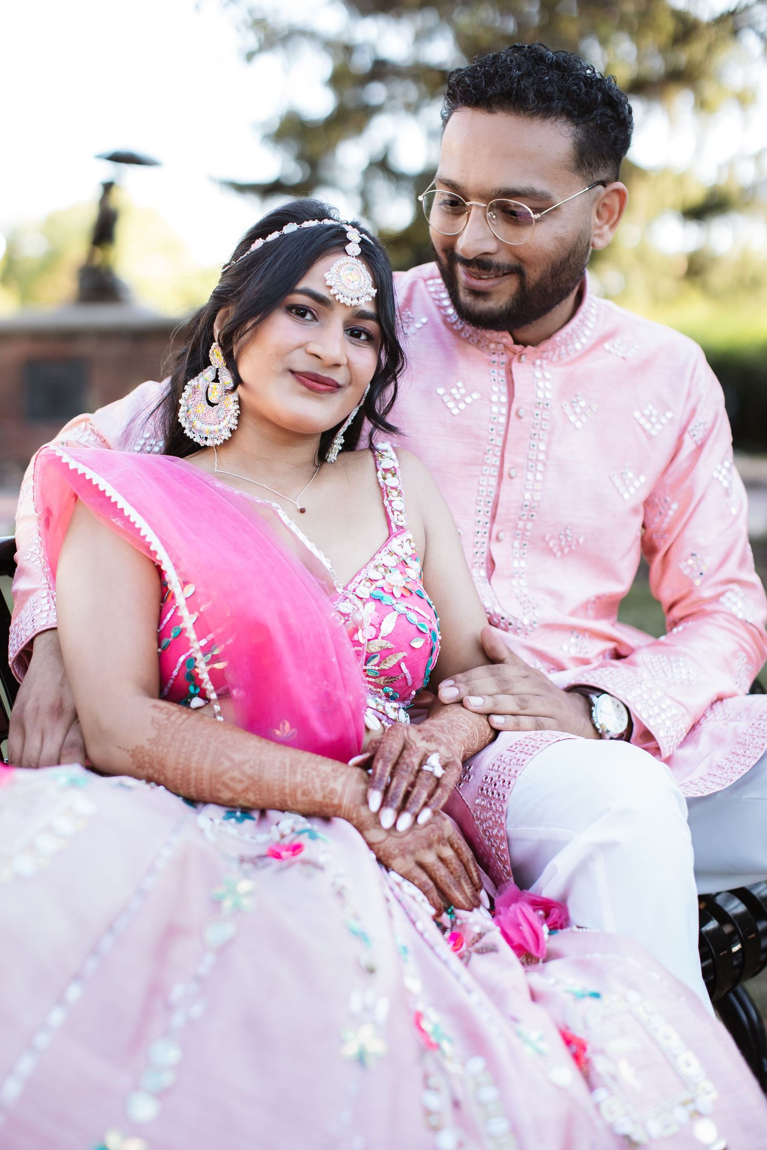 Indian couple in pink traditional wedding attire sitting together outdoors smiling.