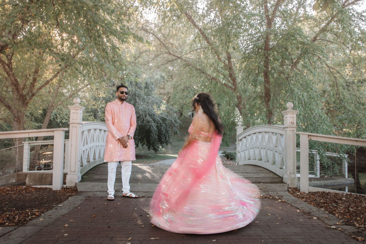 Indian couple in matching pink outfits posing near white ornamental garden bridge.