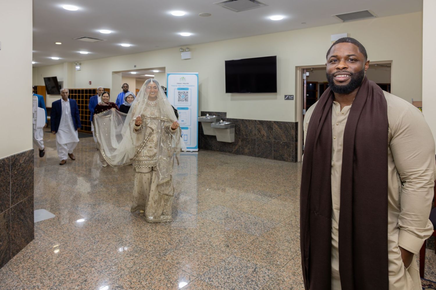 A groom in cream attire smiles as his veiled bride walks toward him in a mosque hallway.