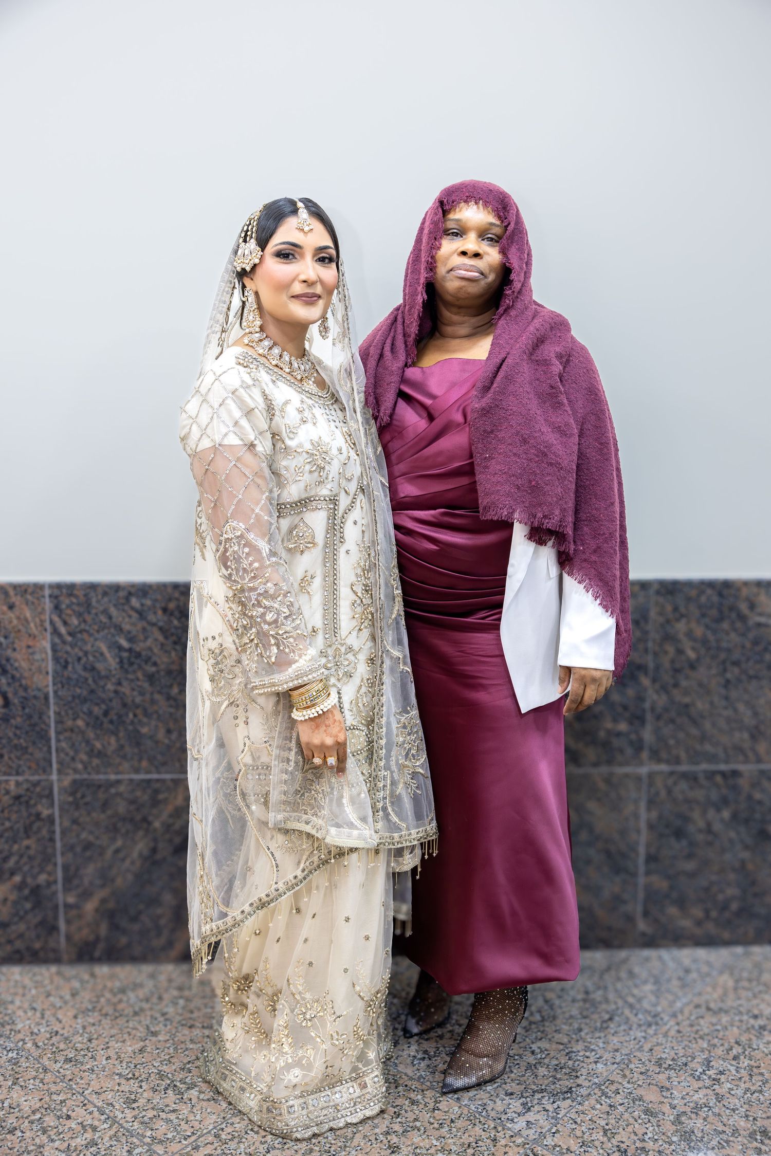 A bride in white embroidered South Asian bridal dress poses closely with a woman in a burgundy gown and headscarf.